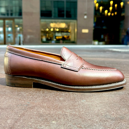The Alden Madison D5211 - Brown CXL Flex Penny Loafer with a classic leather sole is shown outdoors on a flat surface, with softly blurred city lights and buildings in the background.