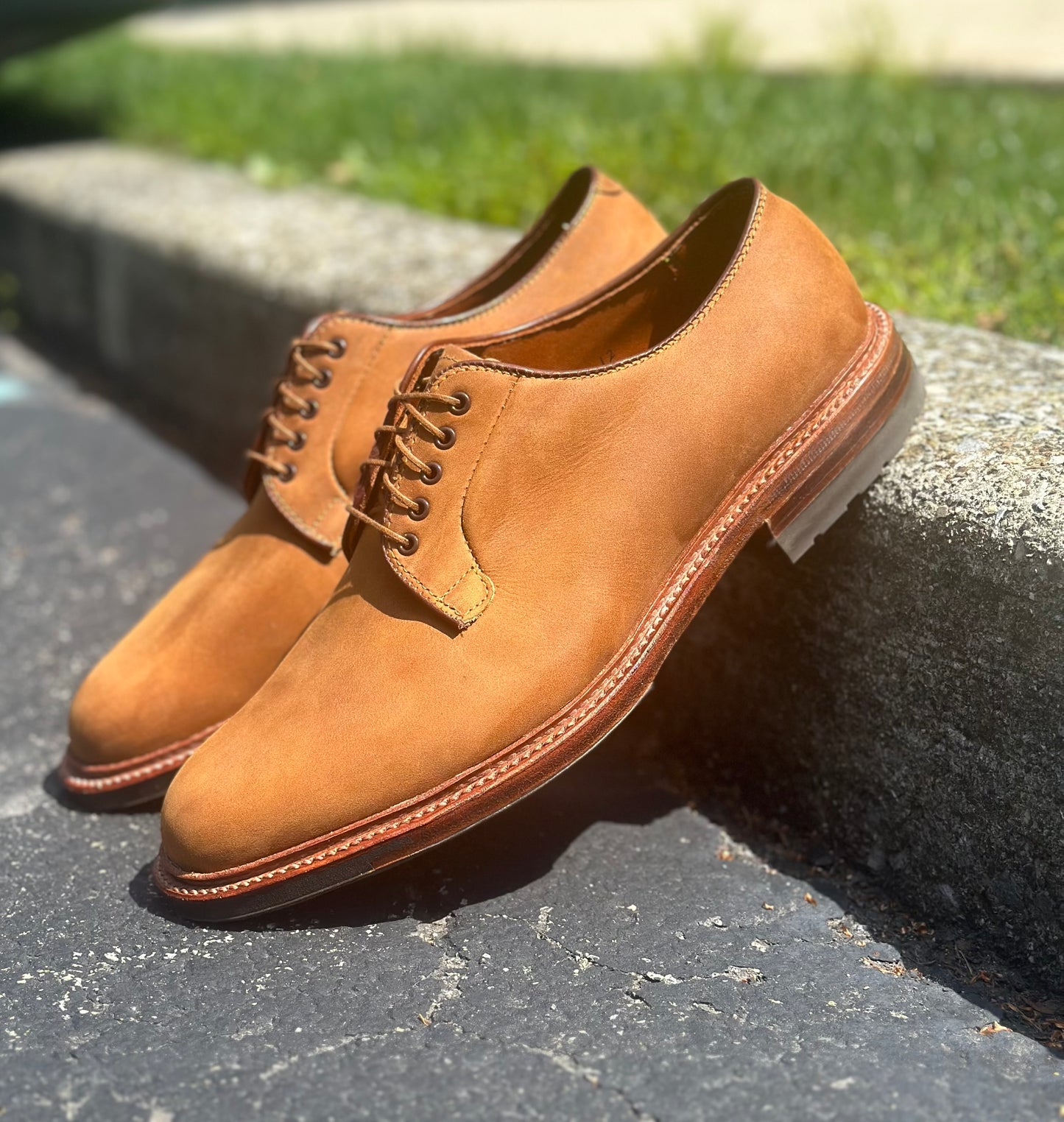 The Alden Madison 9475C Plain Toe Blucher in Tan Smooth Chamois, featuring sturdy Commando soles and laces, rest on a sunlit curb casting shadows on dark pavement, with grass and concrete blurred in the background.