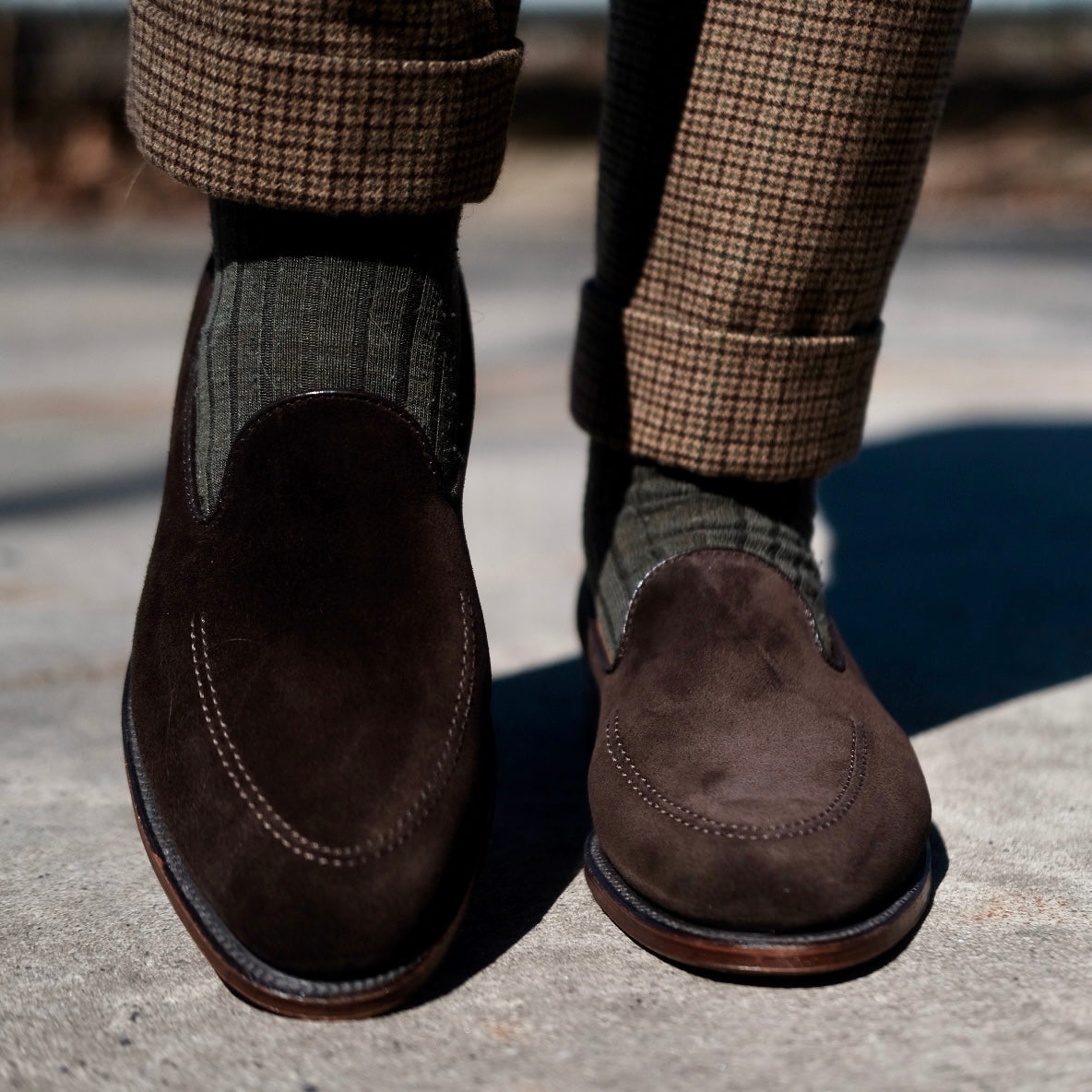 Close-up of a person wearing the ALDEN SHOES D5105 - Vanderbilt in Dark Brown Suede with a Flex Leather Sole, styled with olive green ribbed socks and brown plaid cuffed trousers, standing on concrete.