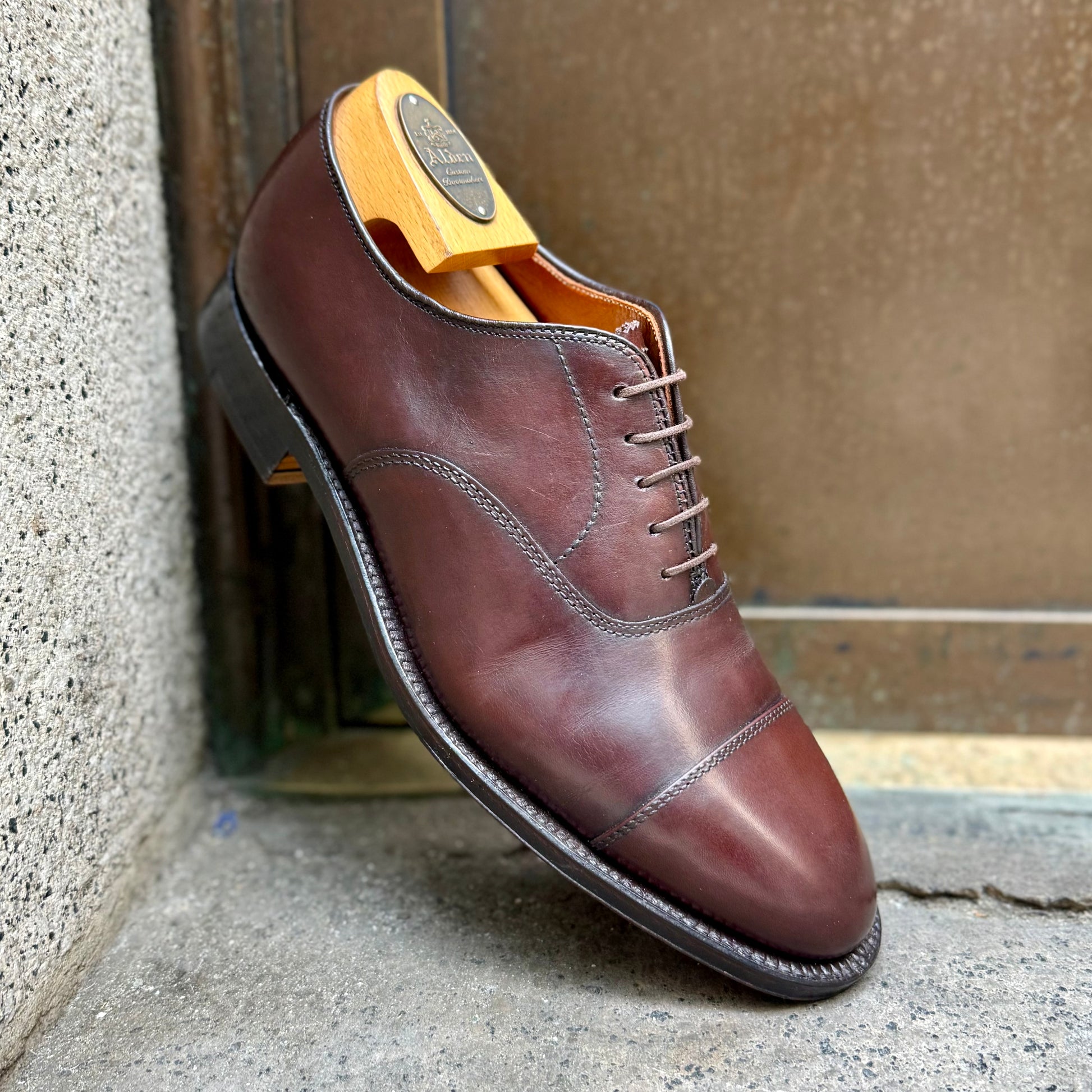 The Alden Madison 920 - Cap Toe Bal in Brown Calfskin, shown with a wooden shoe tree and single oak leather sole on the Hampton last, leans against a concrete and metal surface.