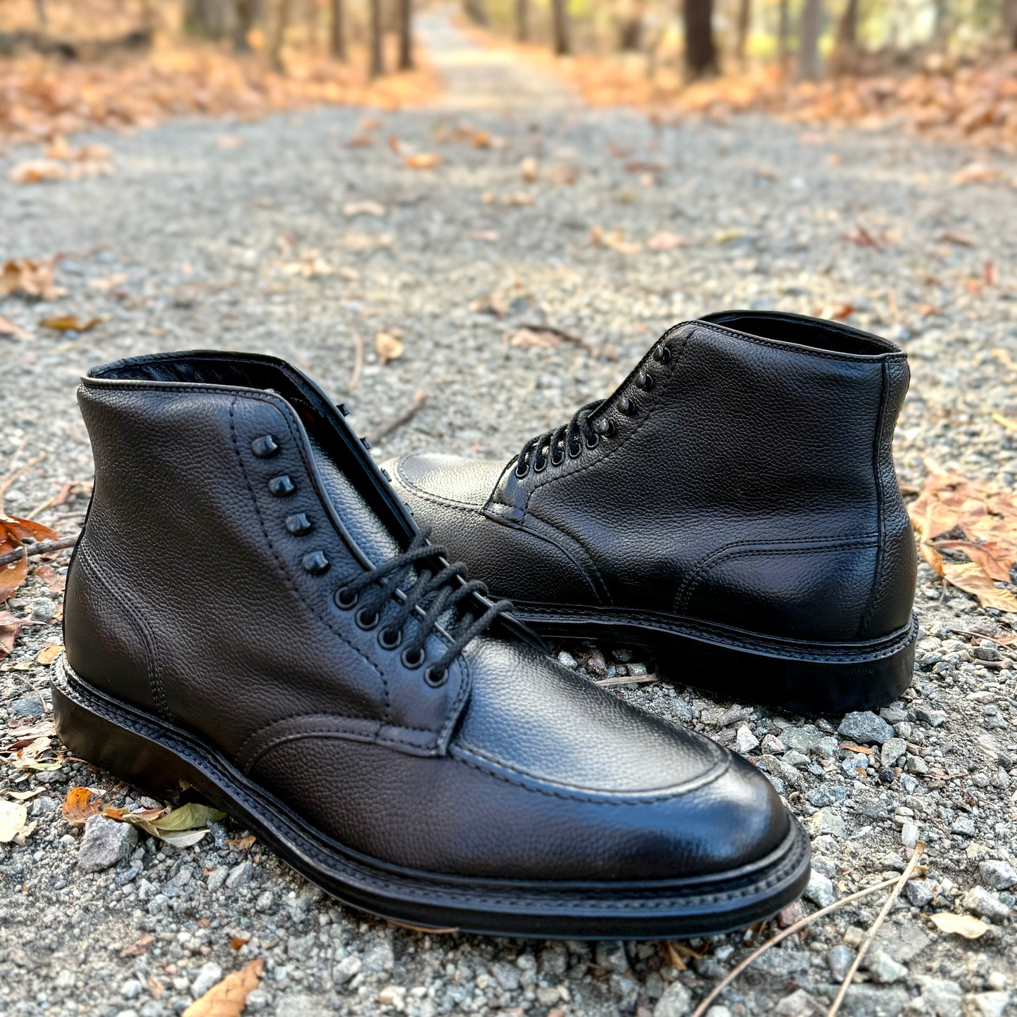 Alden Madison’s D6956LH - The “Han” Boot in Black JS Grain rests on a gravel path with autumn leaves, while a tree-lined walkway fades into the background.