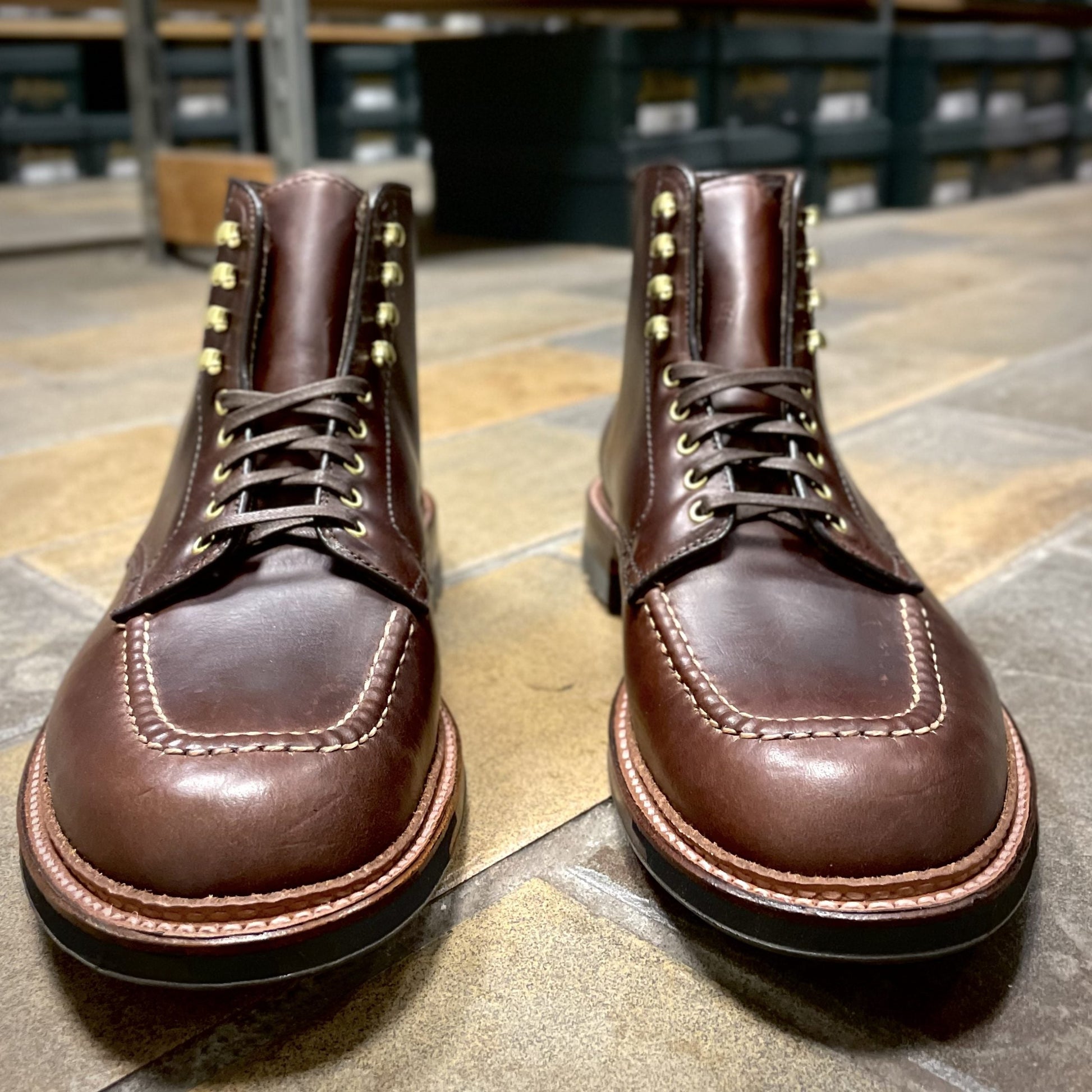A pair of Alden Madisons D1932HC Dirty Bones Indy Boots in brown Chromexcel with gold eyelets and intricate stitching rests on a stone-tiled floor, while blurred shelves with storage boxes form the backdrop.