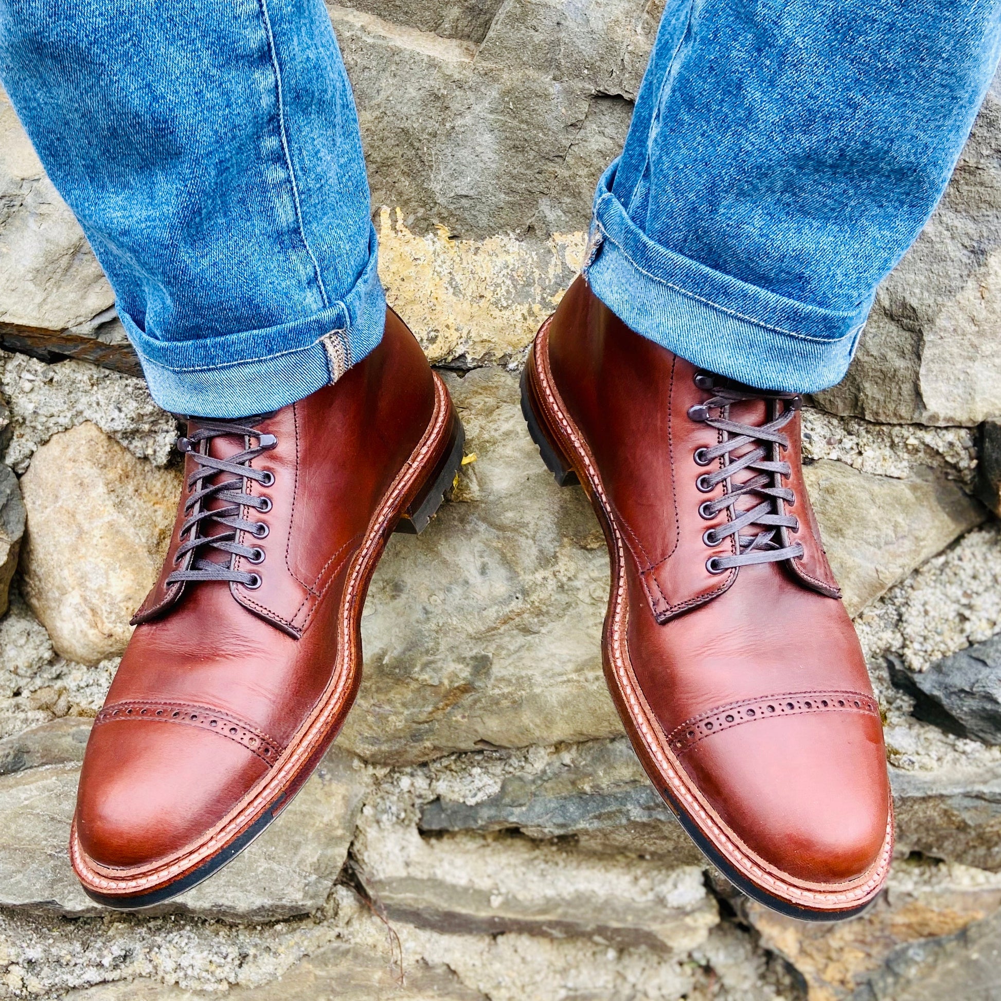 A person wearing rolled-up blue jeans and Alden Madisons 44924HC Jumper Boot in Brown Chromexcel with cap toes and speed hooks stands on a stone surface.