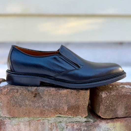 The ALDEN SHOES D2406L "Double Gore" Slip On, a black calfskin ladies’ shoe with a low heel and rubber sole, is shown on a brick surface with a blurred background.
