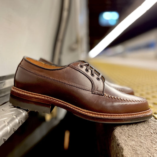 The Alden Madison D1611C - Ranger Mocc in Brown CXL, featuring laces, an antique edge, and detailed stitching, rests on a metallic surface near a train platform edge amid a blurred background.