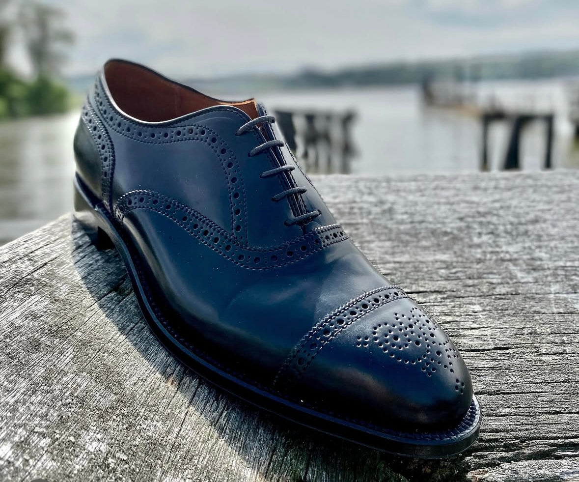 A close-up of the Alden Madison 9841 - Medallion Cap Toe Bal in Black Shell Cordovan, featuring brogue detailing and a refined leather sole, set on a weathered wooden surface near water and docks.