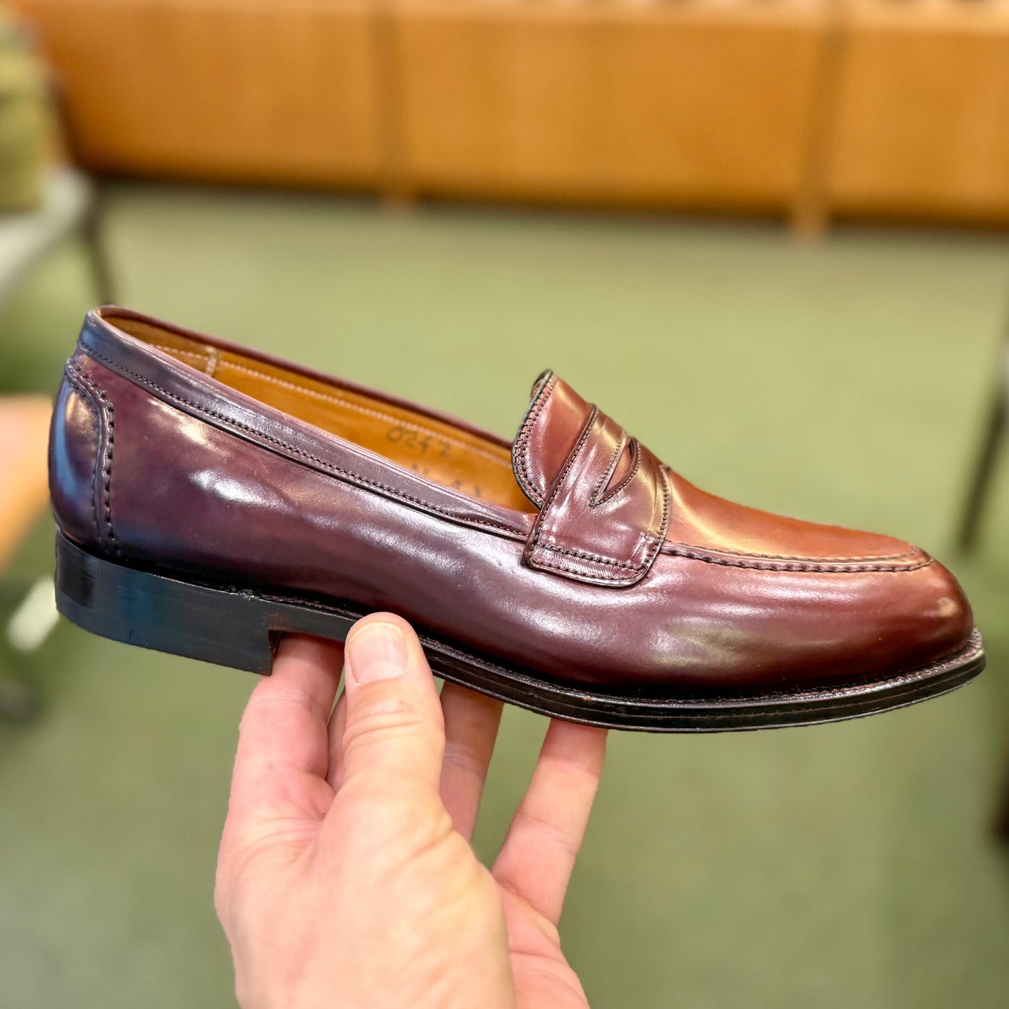 A hand holds the ALDEN SHOES 5750 - Higher Vamp "Midtown" Slip On in Color 8, a polished burgundy loafer with moc toe and penny strap, set against a blurred indoor backdrop with green flooring and brown furniture.