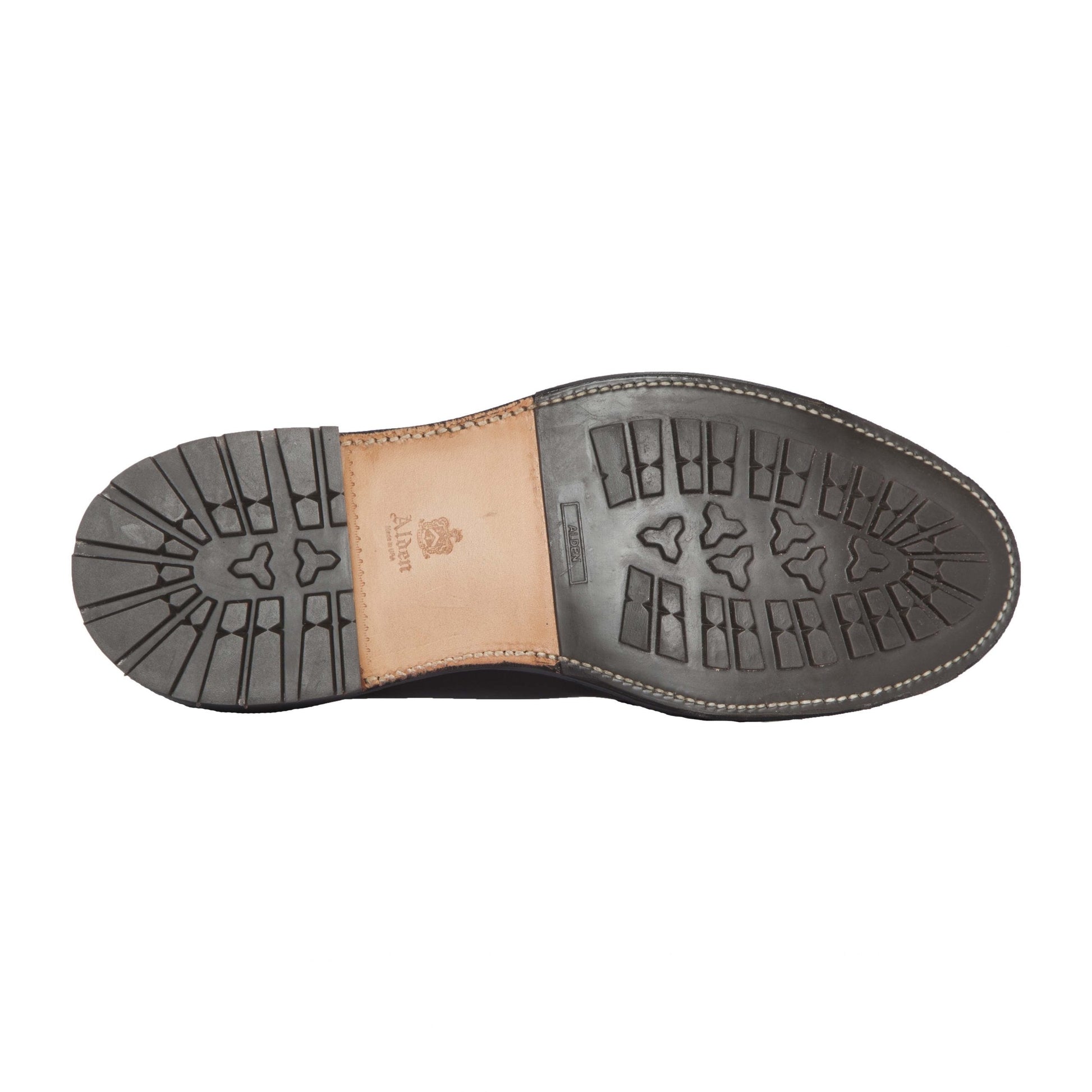 The image shows the 9751C Long Wing Blucher by Alden Madison with a black commando sole featuring triangular tread patterns and horizontal lines for better grip. A tan leather section near the heel displays embossed branding inspired by Black Shell Cordovan craftsmanship.