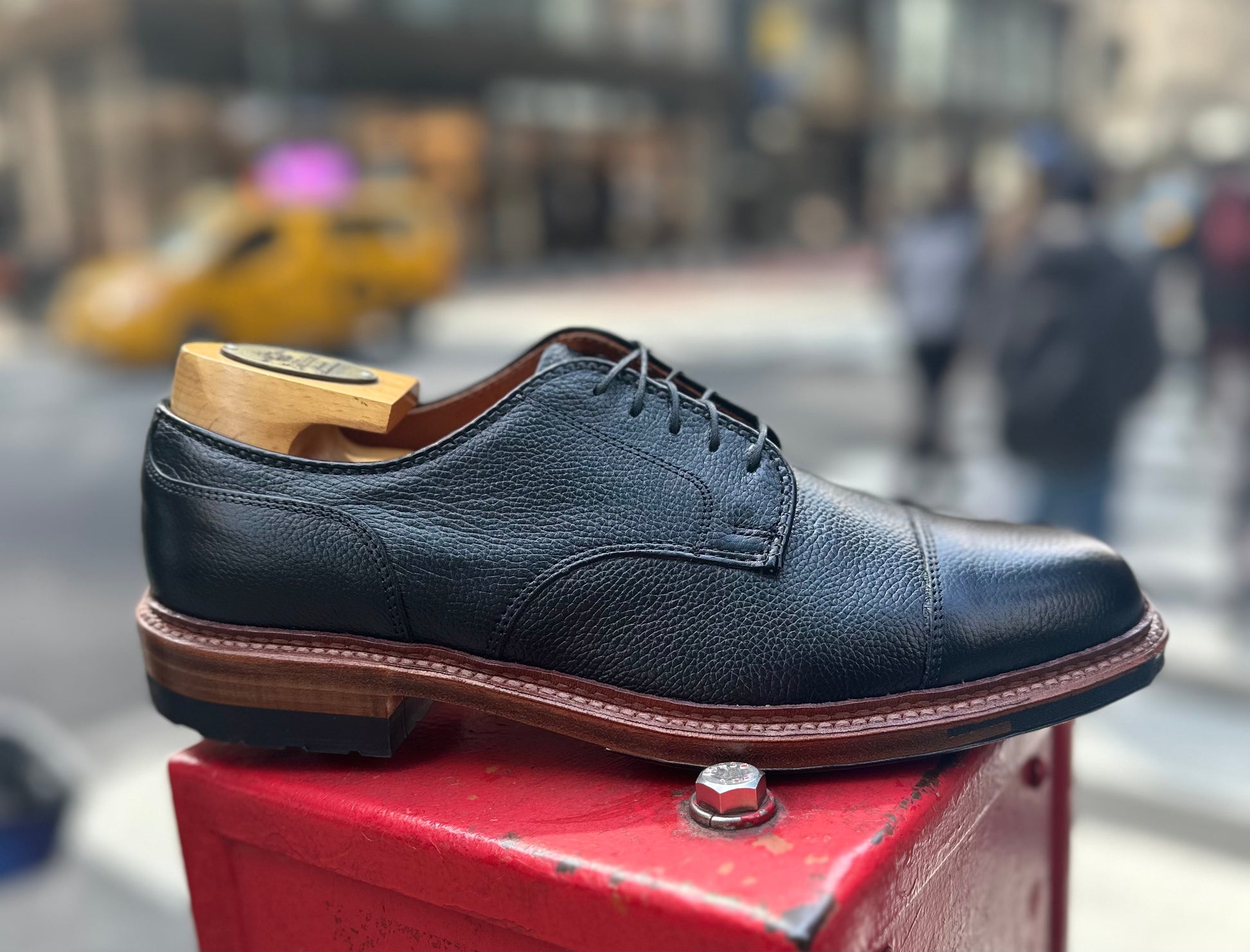 The Alden Madison 22826C - Black JS Grain "Derosa" Blucher, paired with a wooden shoe tree, is shown outdoors on a red metal surface with a blurred city street and people in the background.