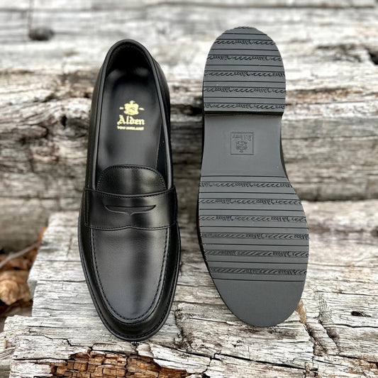 A pair of ALDEN SHOES D5219L Penny Loafers in black lady calf with rubber soles are displayed outdoors on weathered wooden planks, one upright showing the top and the other flipped to reveal its sturdy sole.