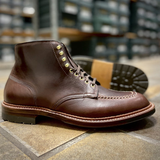 Alden Madisons D1932HC - Dirty Bones Indy Boot in Brown Chromexcel is showcased with stitching details and brass eyelets. One boot stands upright, and the other on its side, showing the Trubalance Last sole against a textured floor with shoe box shelves in the backdrop.