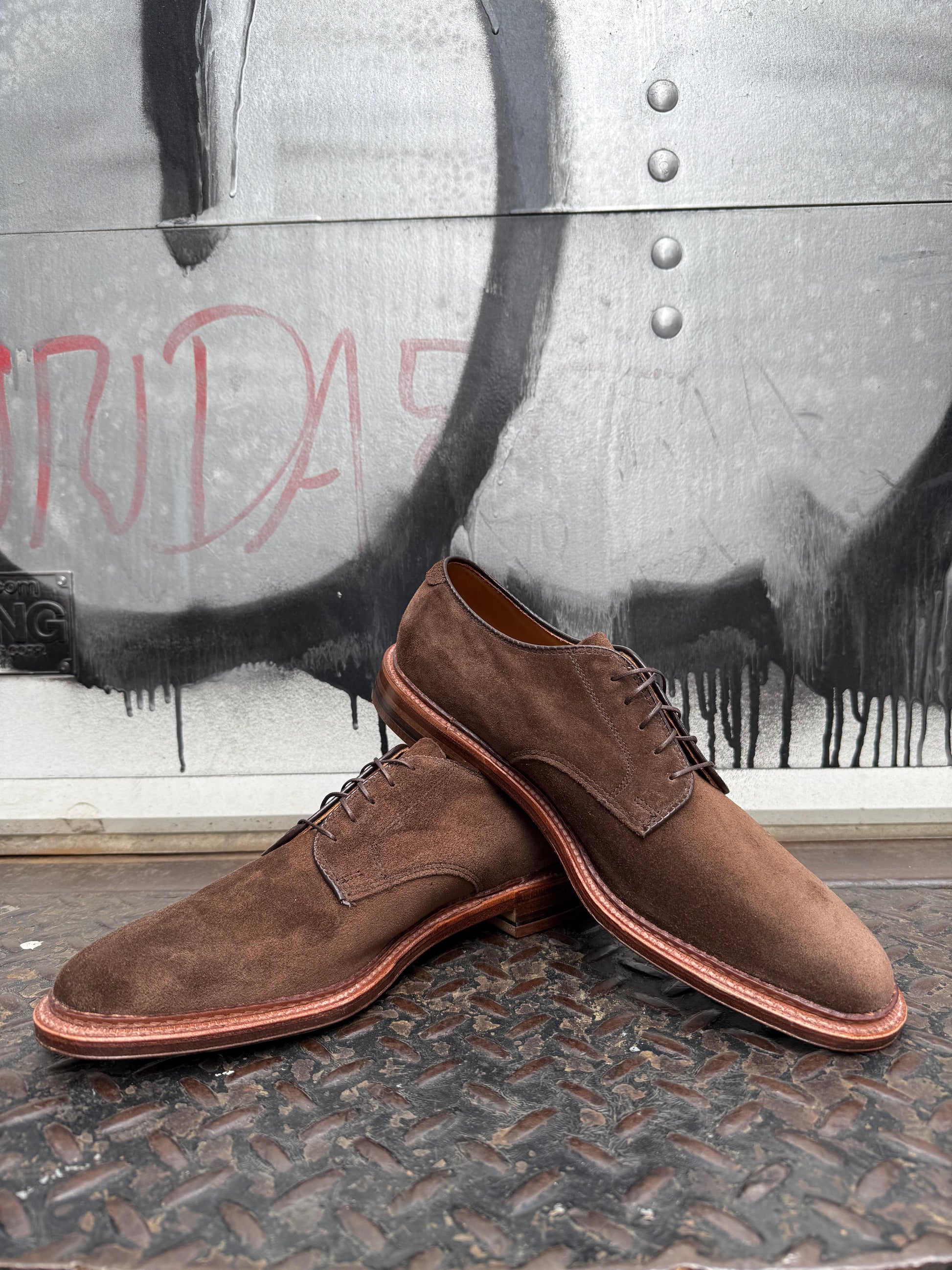 The Alden Madison 29335F - Unlined Dover in Brown Suede dress shoes with Flex Leather Soles are shown outdoors on a metal surface, set against a graffitied wall with black and red markings.