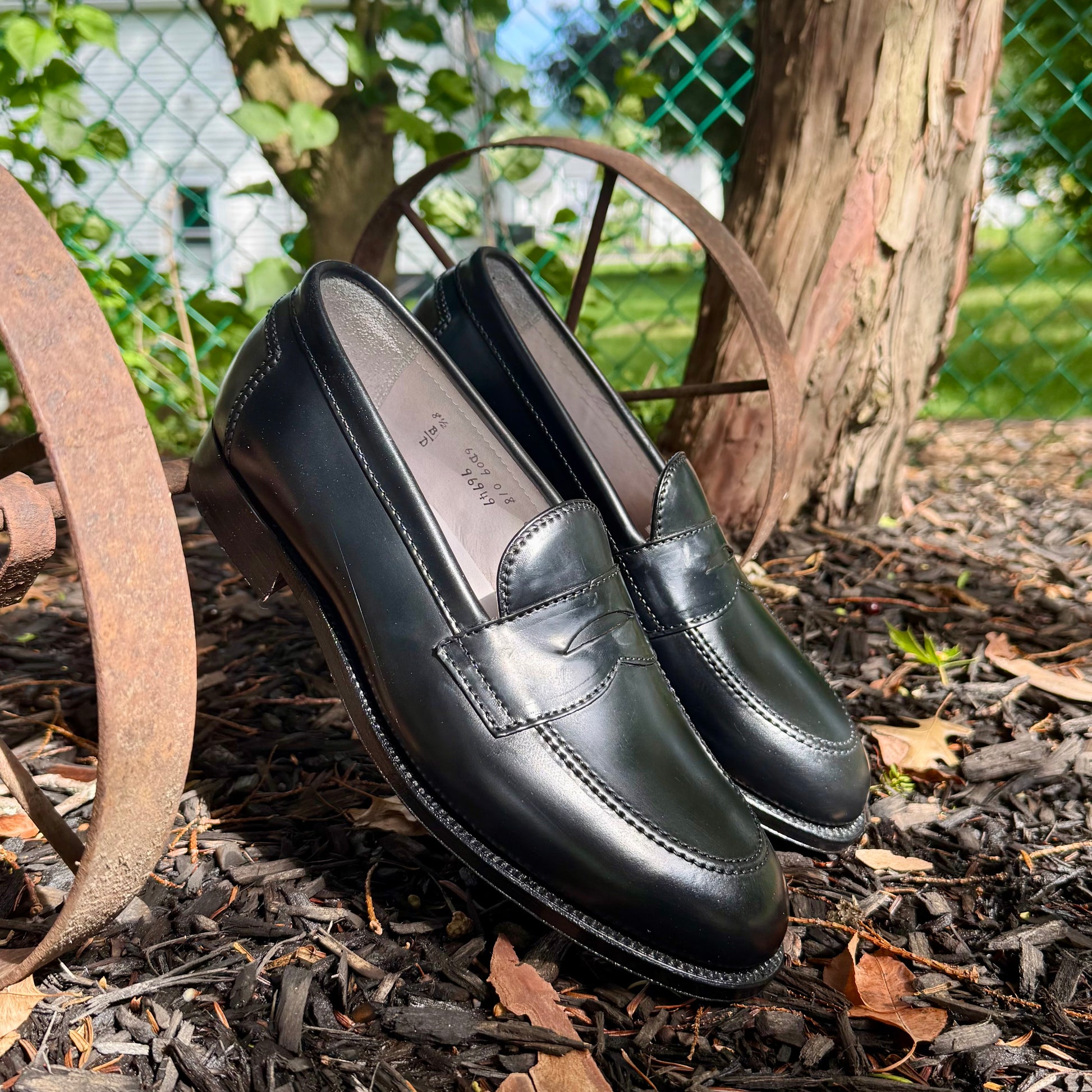A pair of Alden Madison 96949 - Copley Penny Loafers in Black Shell Cordovan, crafted on the Copley Last with a sleek 270 Flat Welt, rests on old metal wagon wheels outdoors, surrounded by mulch, green plants, and sunlight.