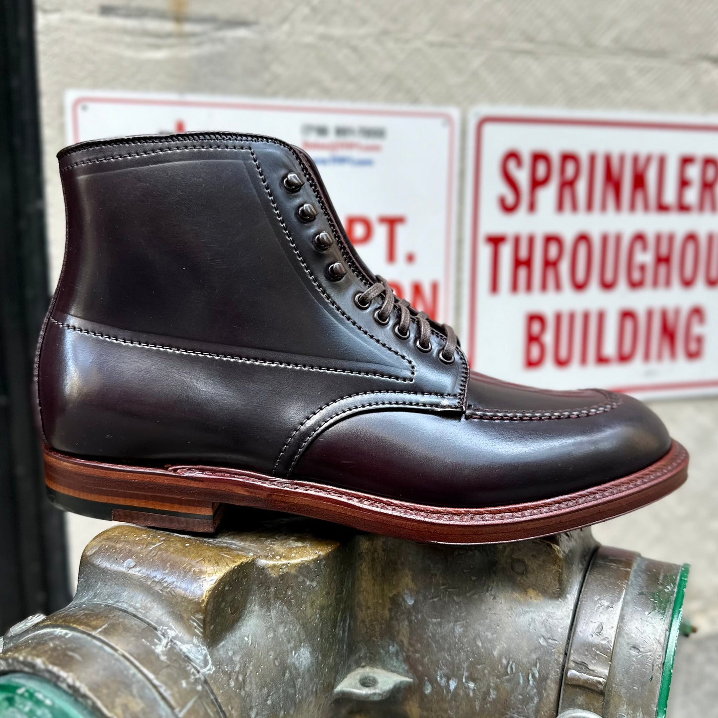 An Alden Madison D2936H - Color 8 Shell Indy Boot, crafted from dark brown leather with detailed stitching and a Double waterlock sole, is displayed outdoors on a metal fixture, with blurred white and red warning signs in the background.