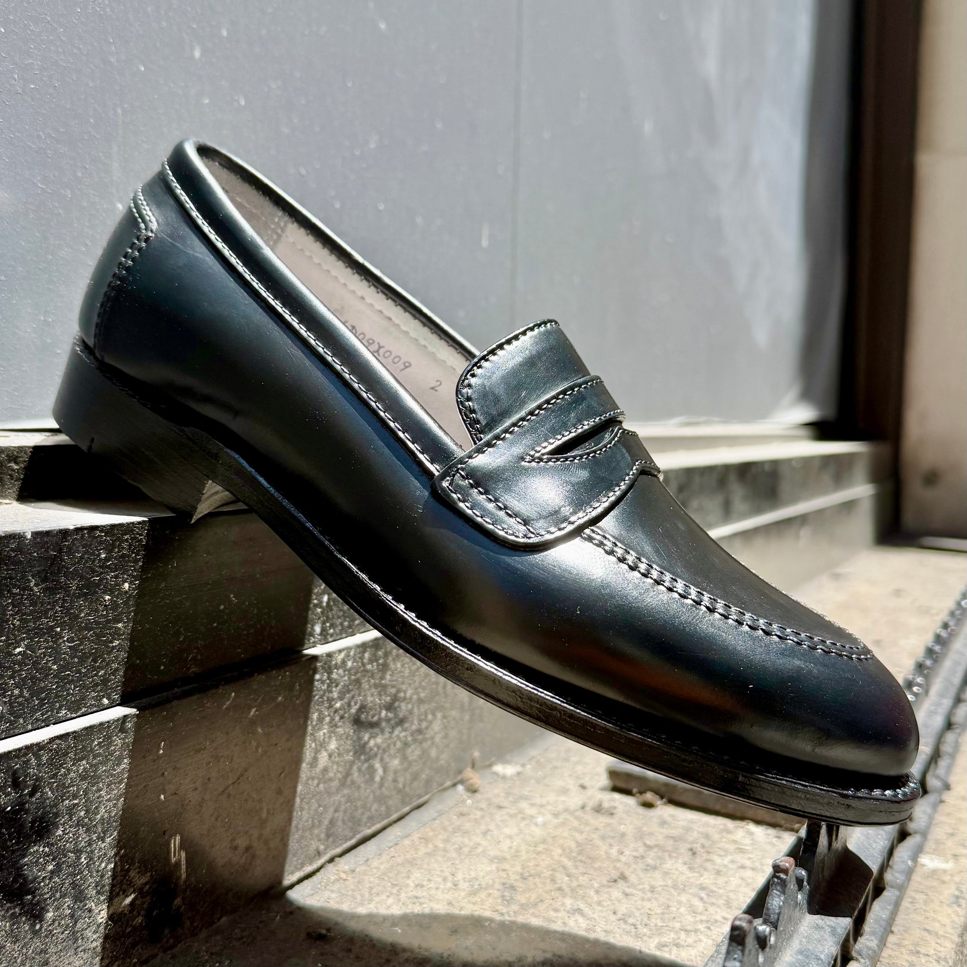 The ALDEN SHOES 5751 High Vamp Loafer in Black Shell Cordovan, crafted on the elegant Aberdeen Last, is shown outdoors on a stone step with sunlight highlighting its polished finish and intricate stitching.