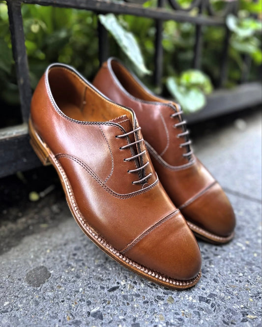 A pair of ALDEN SHOES D5310 - Straight Tip Oxford in Madison Brown Calf is placed on a sidewalk near a black metal fence and green plants.