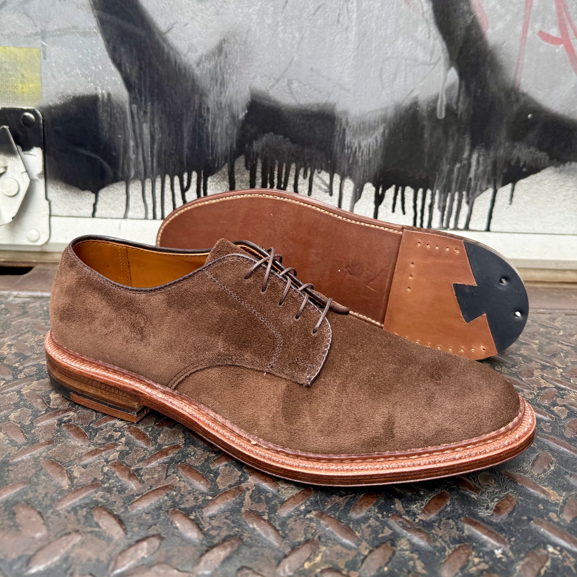 A pair of Alden Madison 29335F - Unlined Dover in Brown Suede shoes, shown on flex leather soles—one upright, one revealing the sole—are displayed on a textured metal surface with graffiti in the background.