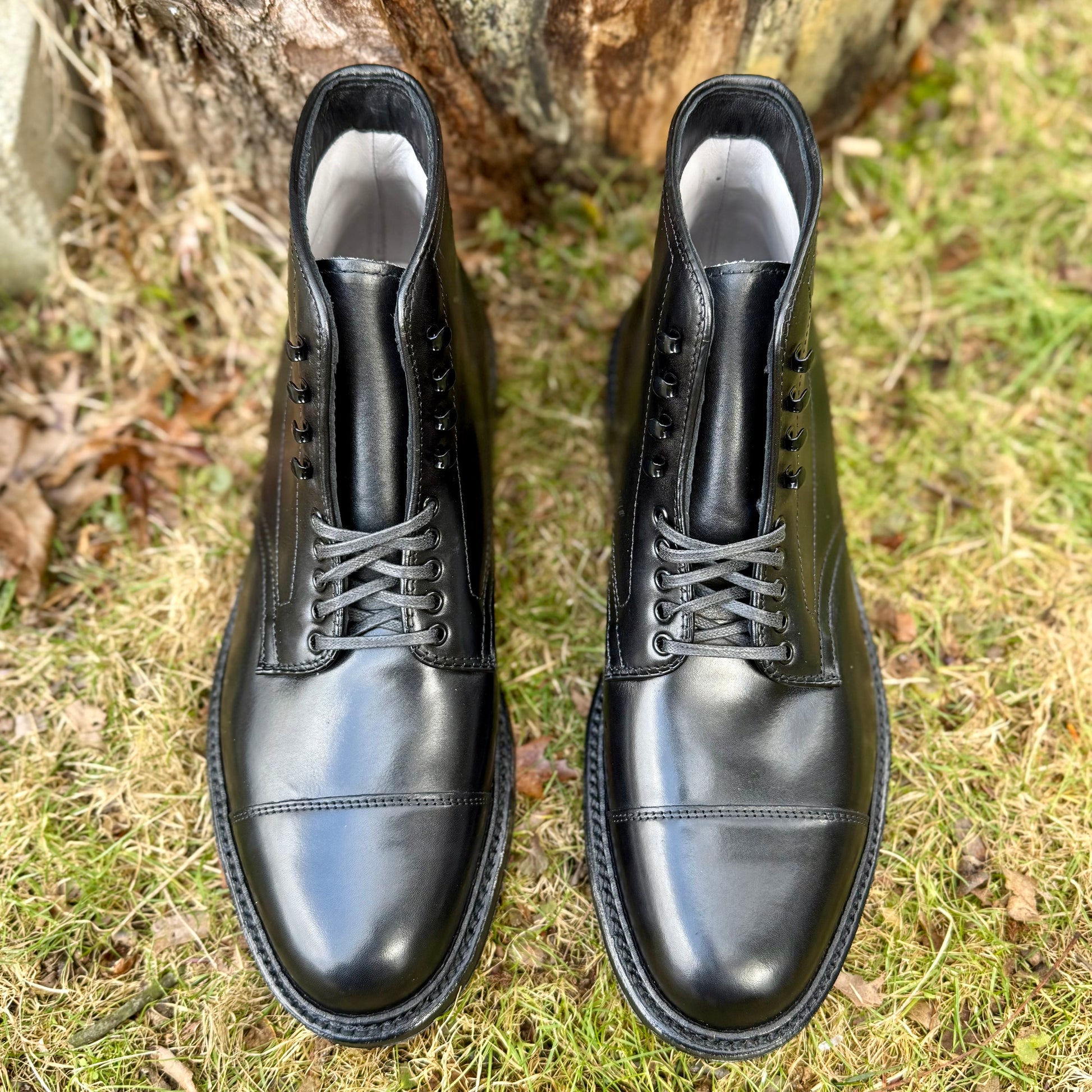 Alden Madison’s 40797HC Black Cap Toe Boot with Commando Sole features glossy black calfskin and laces, displayed outdoors on grass beside a tree trunk and scattered leaves.
