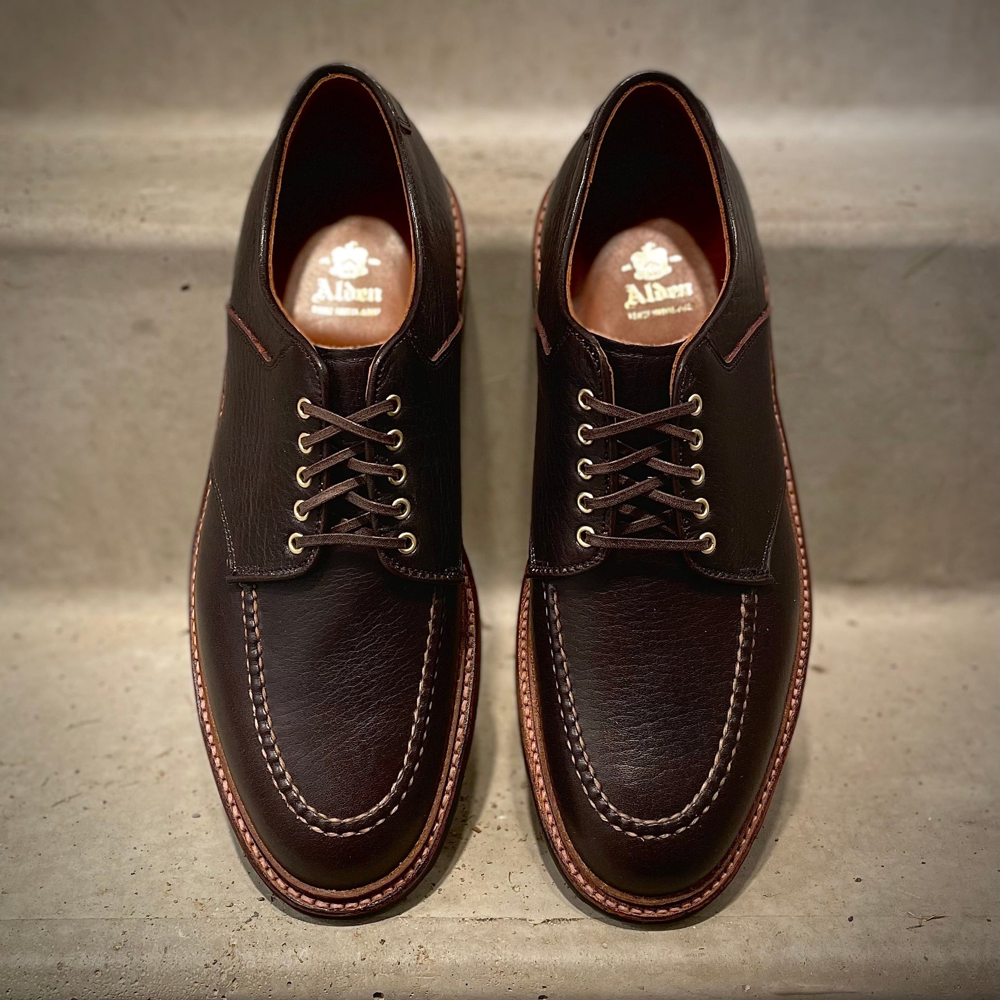 A pair of Alden Madison D1613C The Belmont Saddle Blucher Lux Arabica Calf dress shoes with white stitching and laces, side by side on a light surface. Gold Alden branding on the insoles. Crafted on the Barrie Last for a classic fit.