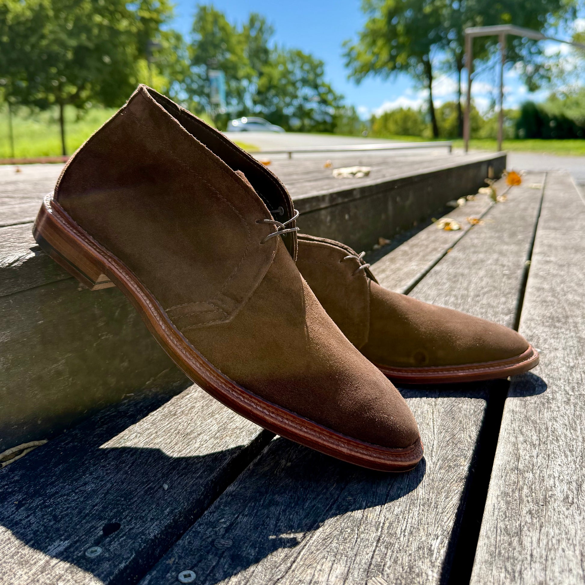 The Alden Madison 1492 - Unlined Chukka in Brown Suede sits on a wooden deck outdoors, sunlight highlighting its Single Flex Leather Sole, with green trees and a clear blue sky in the background.