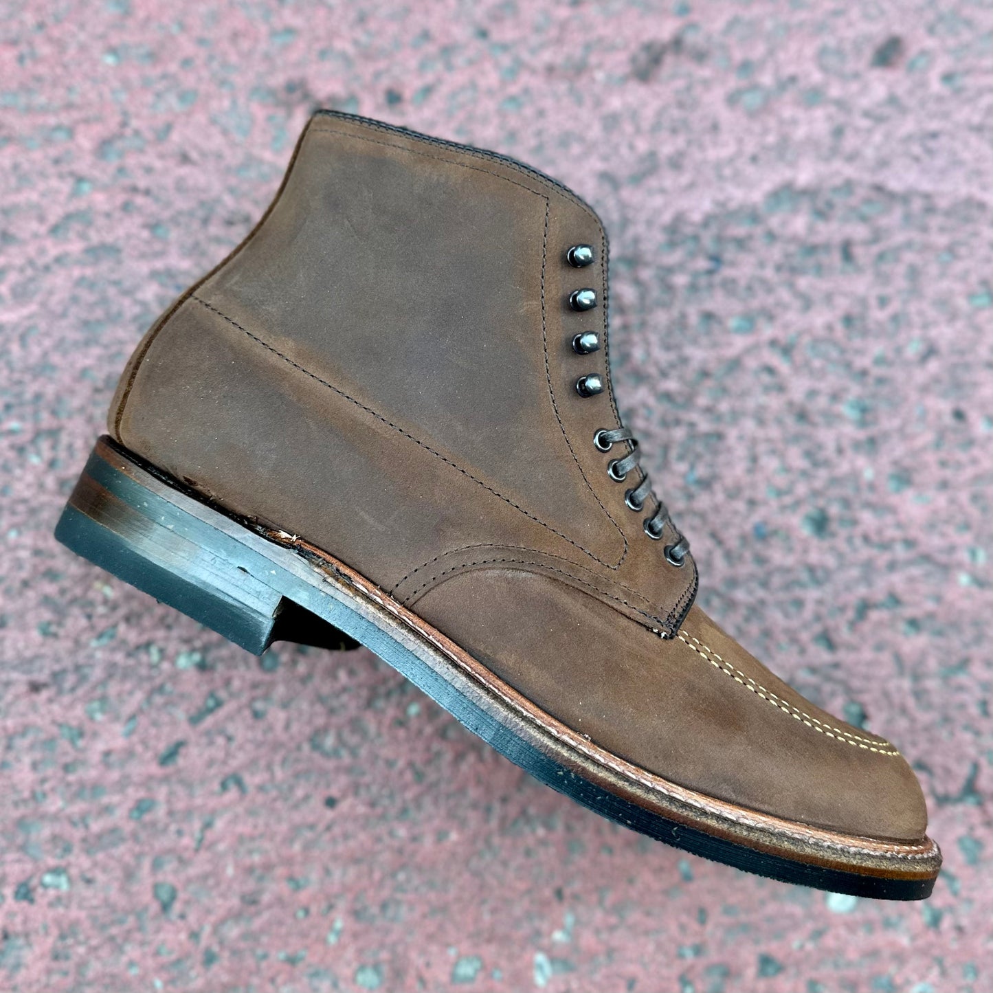 An Alden Madison D2909H - Indy Boot in Tobacco Smooth Chamois with black laces and dark metal eyelets rests on a textured pink surface, crafted on the Trubalance Last, featuring a raised heel and moc toe design with contrast stitching.