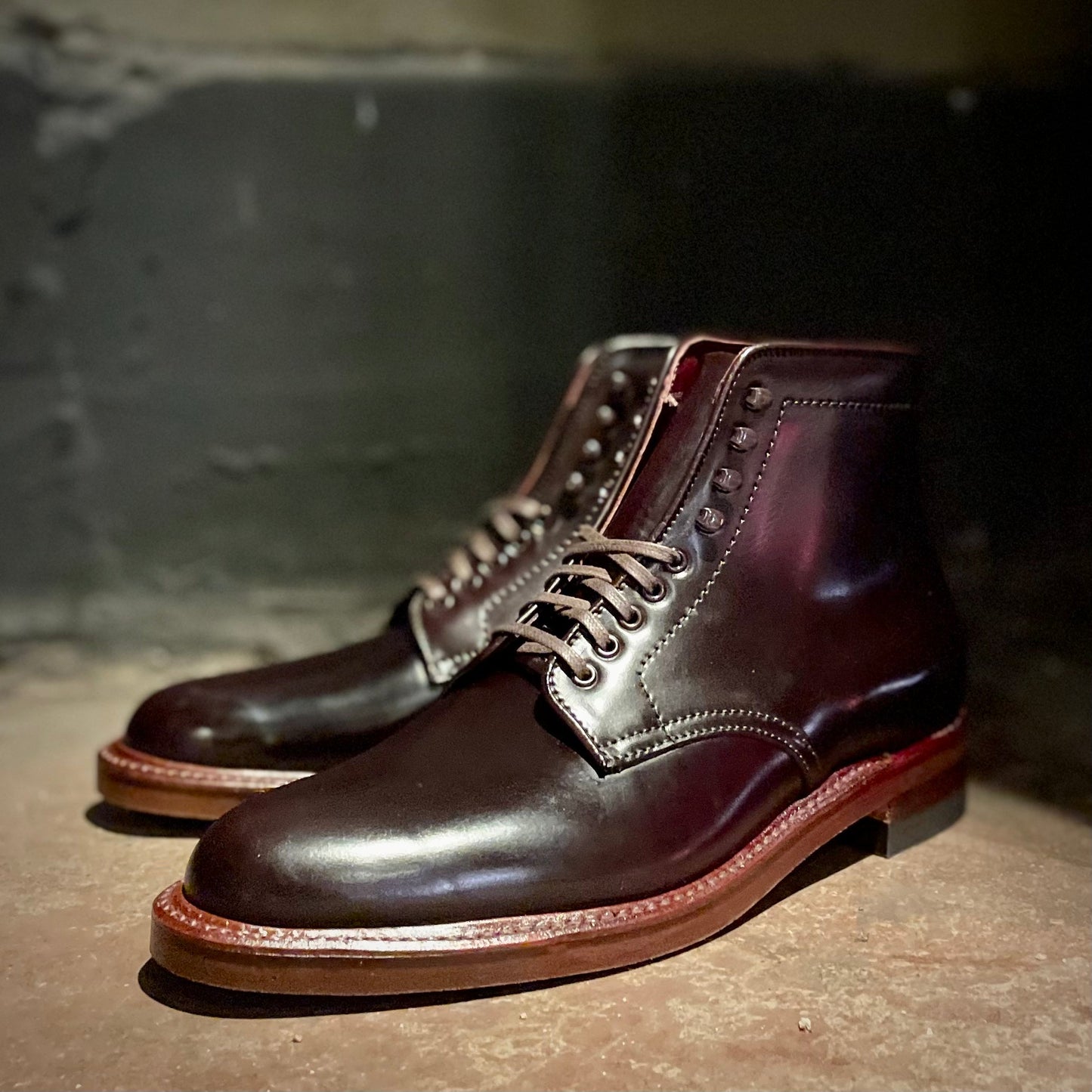 A pair of Alden Madison D1854H - Plain Toe Boots in Color 8 Shell Cordovan, featuring a neocork sole with a rich reddish-brown hue, are displayed on a rustic surface against a dimly lit background.