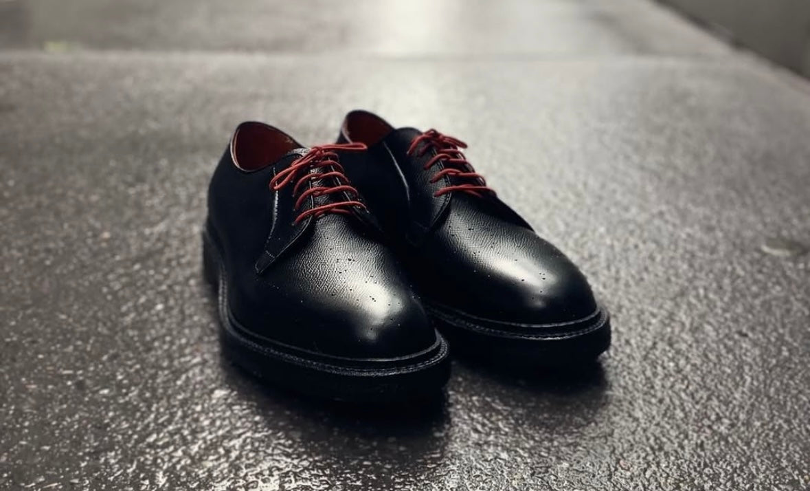 The Alden Madison 949 - All Weather Walker in Black Alpine Grain with red laces rests on wet pavement, water droplets showing recent rain. Featuring the Barrie last and crepe sole, this pair offers both comfort and durability.