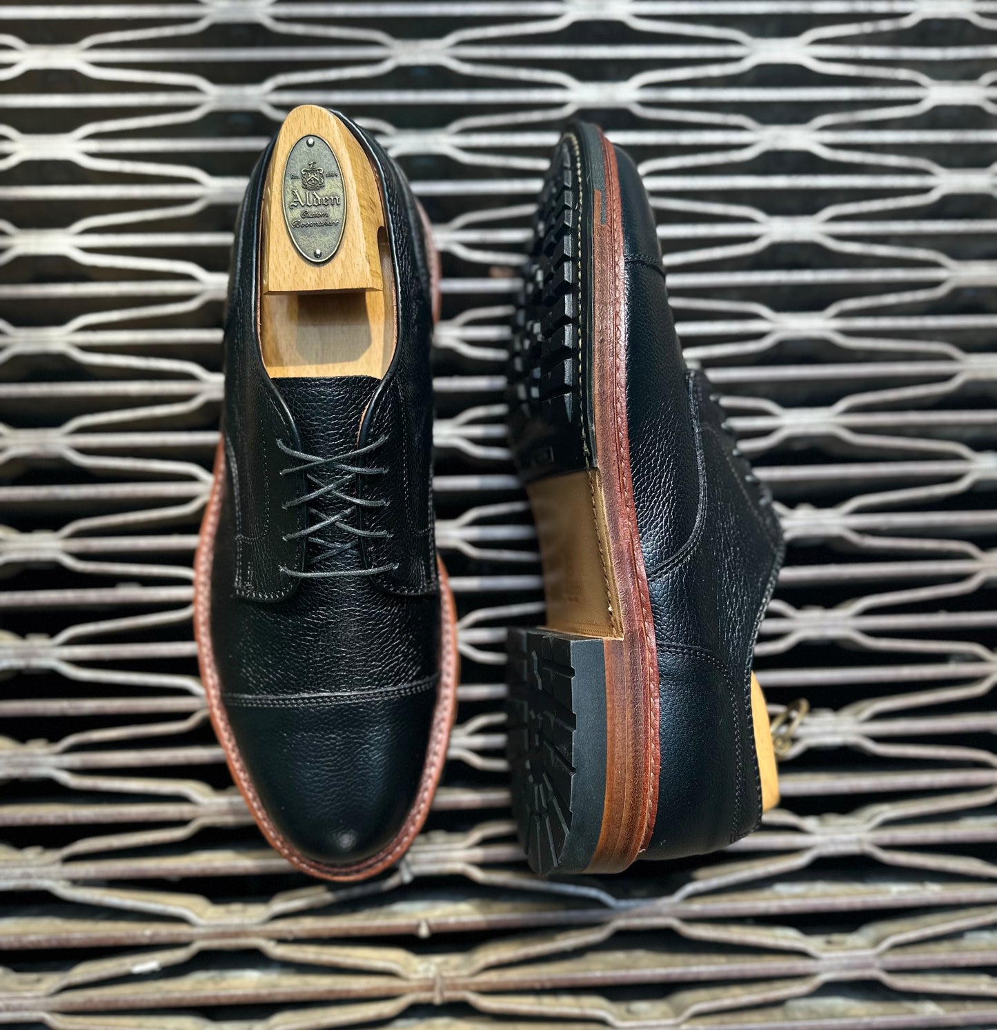 Two Alden Madison 22826C Black JS Grain "Derosa" Blucher shoes with brown Commando soles are shown on a metal grate: one is upright with a wooden shoe tree, the other lies on its side highlighting sole and heel details.