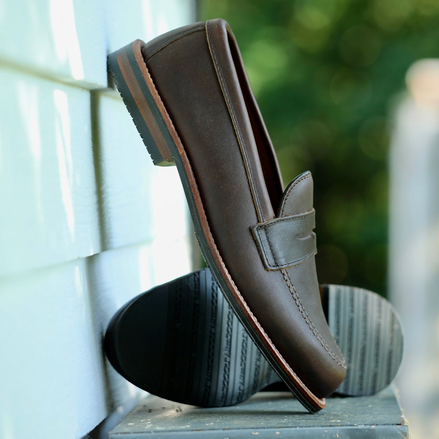 A pair of ALDEN SHOES 5730L- Penny Mocc in Kudu with Rubber Sole, featuring Mocc Toe detailing, are shown outdoors—one upright and one on its side—against a blurred green and white backdrop.