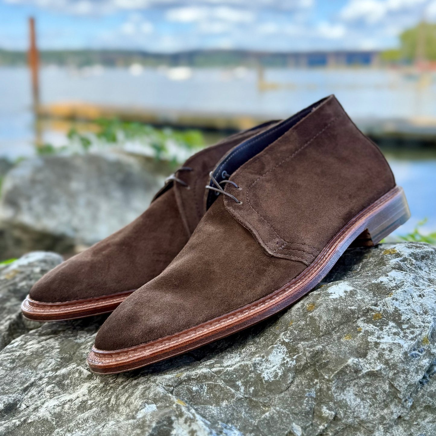 The Alden Madison 1492 - Unlined Chukka in Brown Suede, featuring a single flex leather sole, sits atop a large gray rock with a softly blurred background of water, greenery, and a cloudy sky.