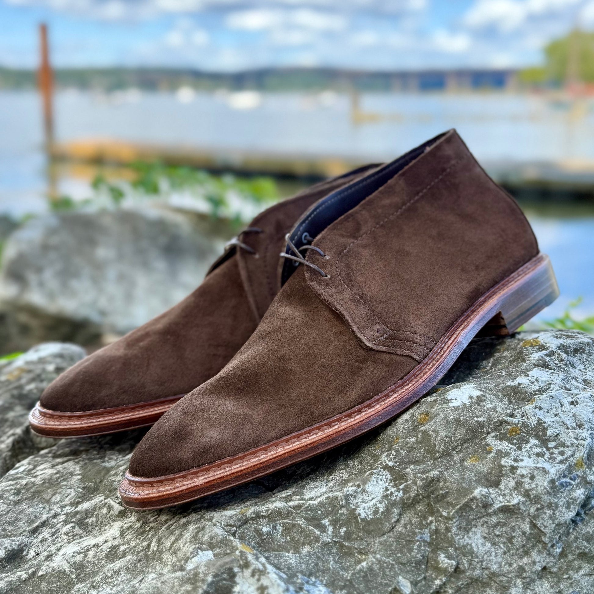 The Alden Madison 1492 - Unlined Chukka in Brown Suede, featuring a single flex leather sole, sits atop a large gray rock with a softly blurred background of water, greenery, and a cloudy sky.