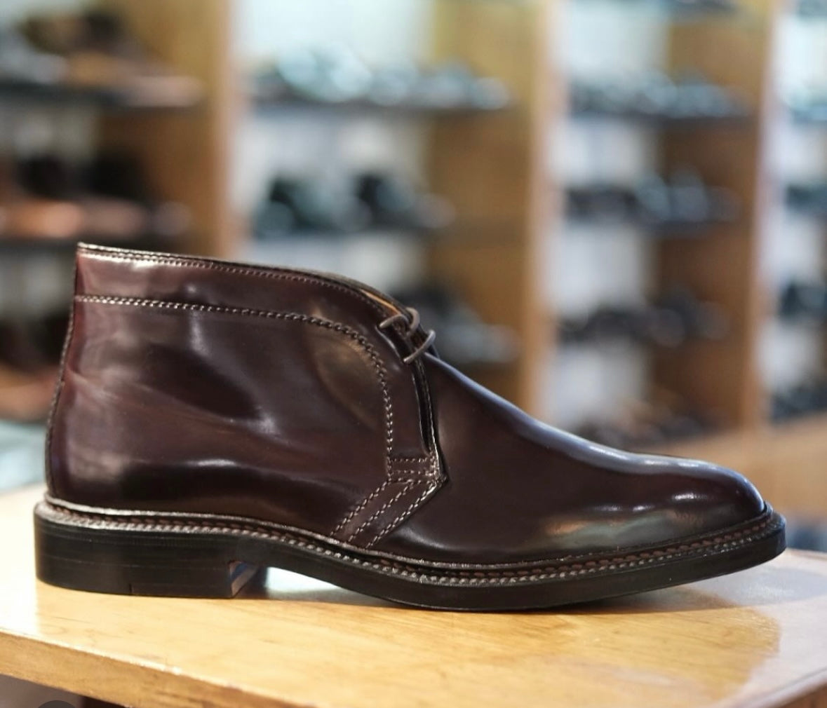 The Alden Madison 1339 - Chukka Boot in Color 8 Shell Cordovan, featuring a Double Oak Leather Sole, is showcased on a wooden surface with blurred shelves of shoes in the background.