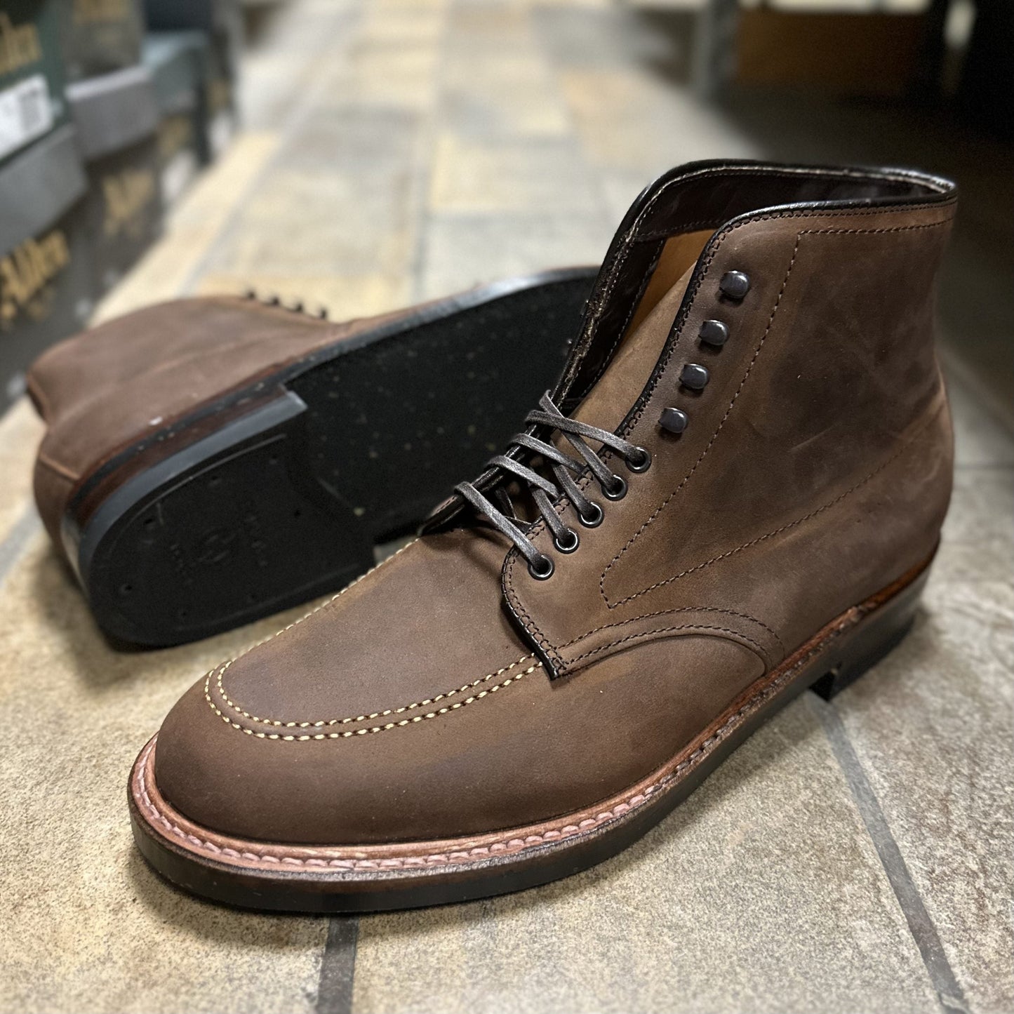 A close-up of Alden Madisons D2909H Indy Boot in Tobacco Smooth Chamois reveals brown leather and dark soles. One stands upright, showcasing its laces and Trubalance Last stitching, while the other lies on its side against a tiled floor and shoe boxes.