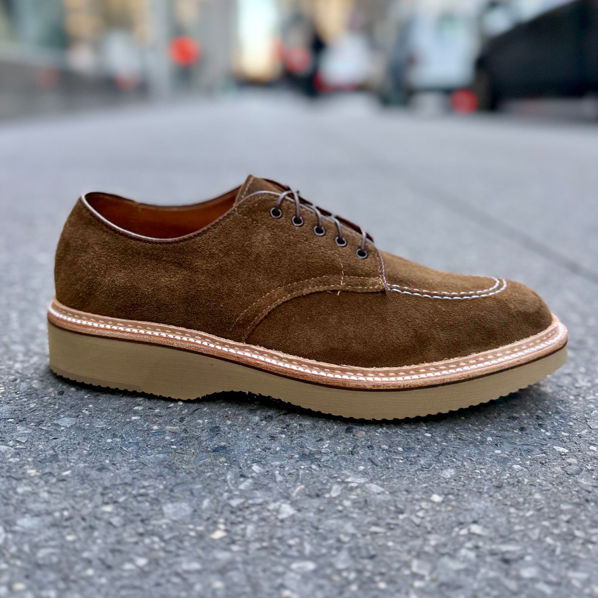 An ALDEN SHOES M7601Y - Snuff Suede Indy Shoe with Wedge, featuring a thick beige sole and white stitching, stands on a concrete sidewalk as the city street blurs in the background.