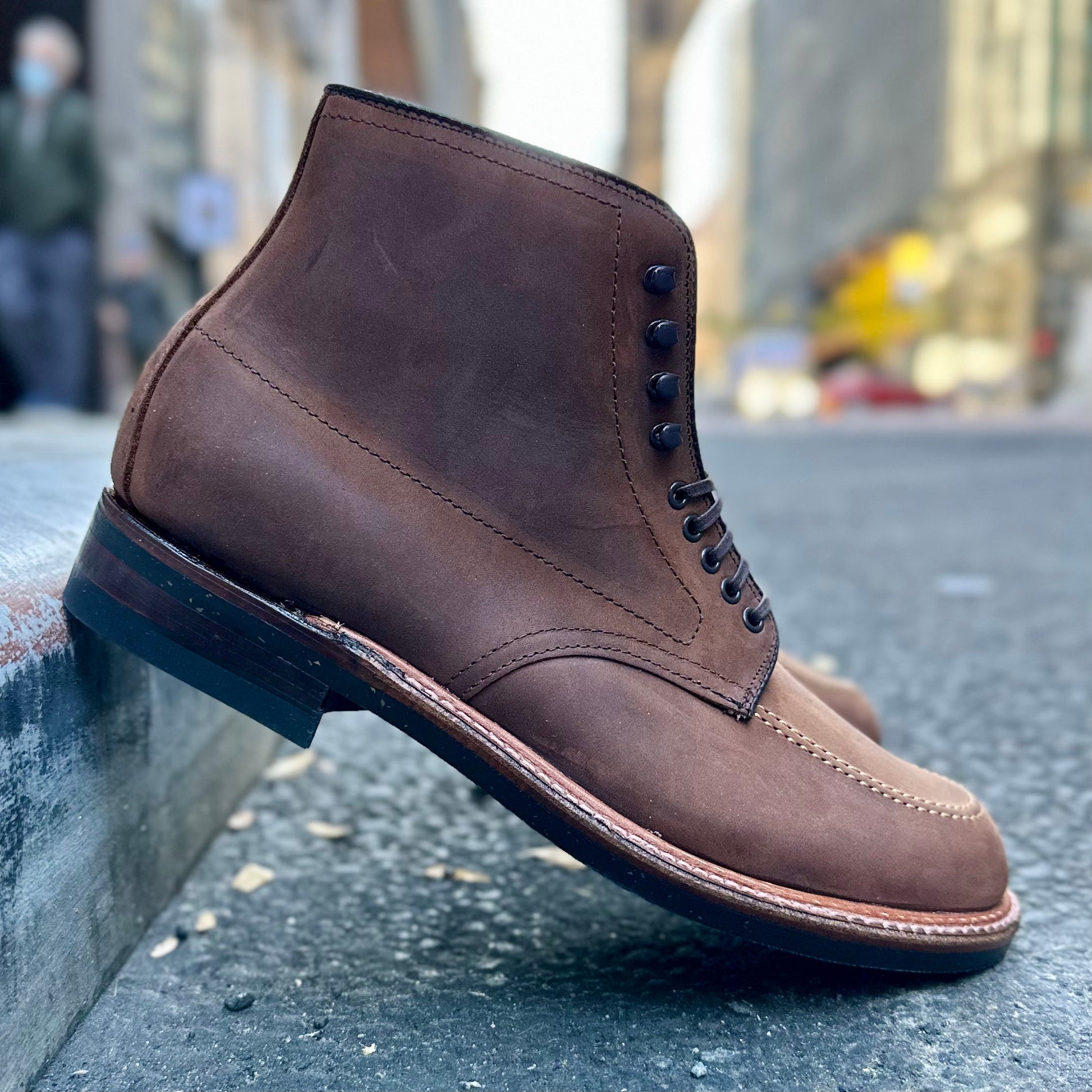 A close-up of the Alden Madison D2909H - Indy Boot in Tobacco Smooth Chamois featuring black laces on concrete, with a blurred urban street in the background highlighting its craftsmanship and refined Trubalance Last design.