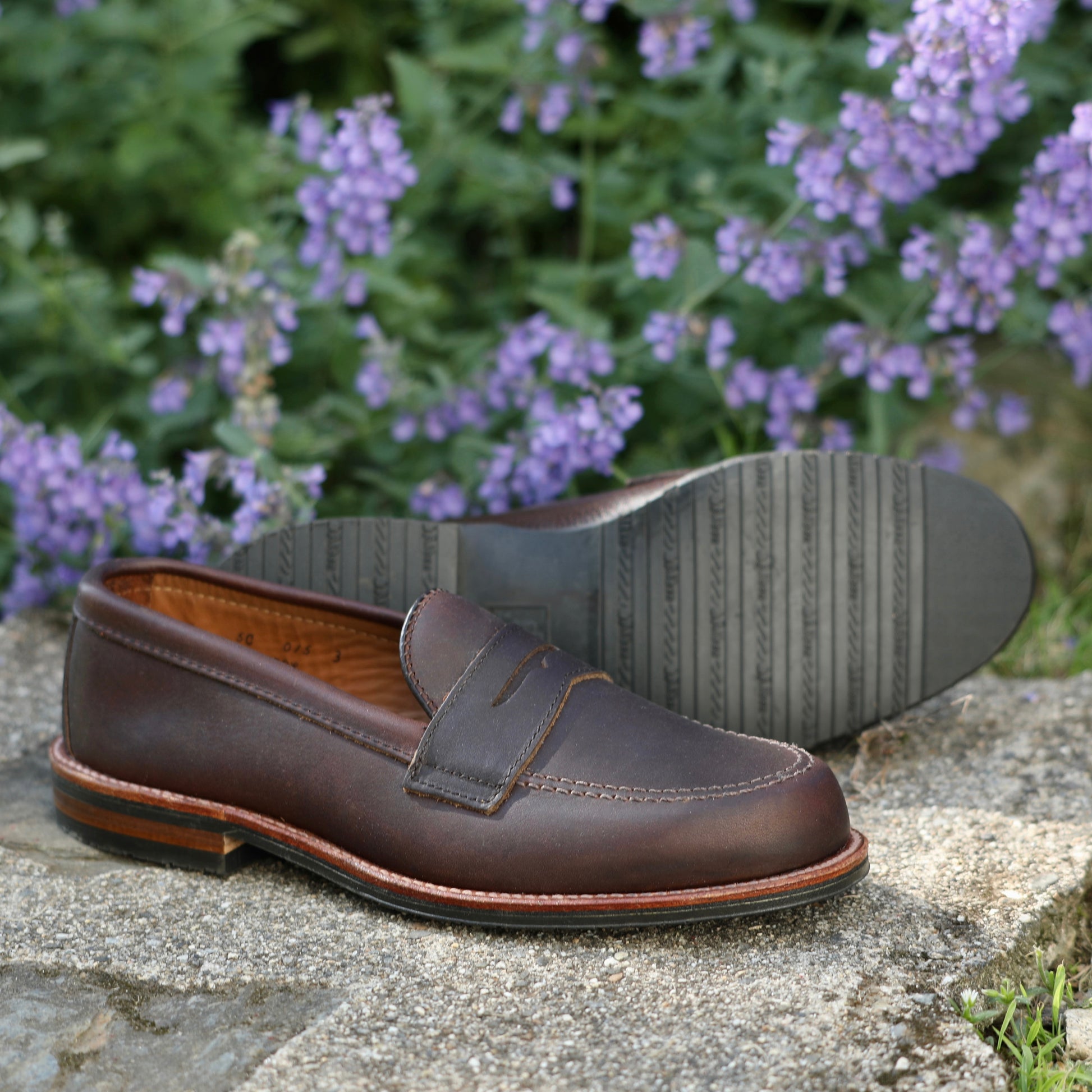 The ALDEN SHOES 5730L-Penny Mocc in Kudu with Rubber Sole is shown outdoors on a stone surface, one shoe upright and the other on its side, with purple flowers and green foliage softly blurred in the background.