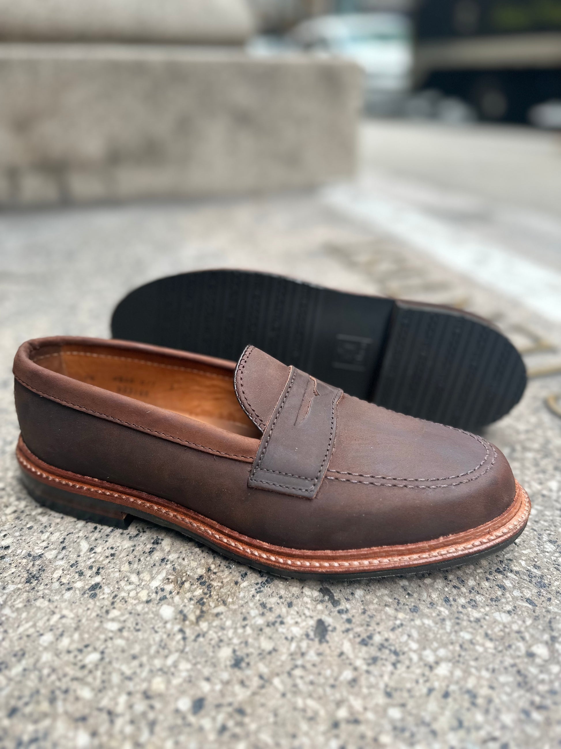 The Alden Madison D2216L - Smooth Tobacco Chamois Penny Loafer, with antique edge and visible stitching on a rubber sole, stands upright on concrete while another lies sideways, revealing its intricate tread pattern.