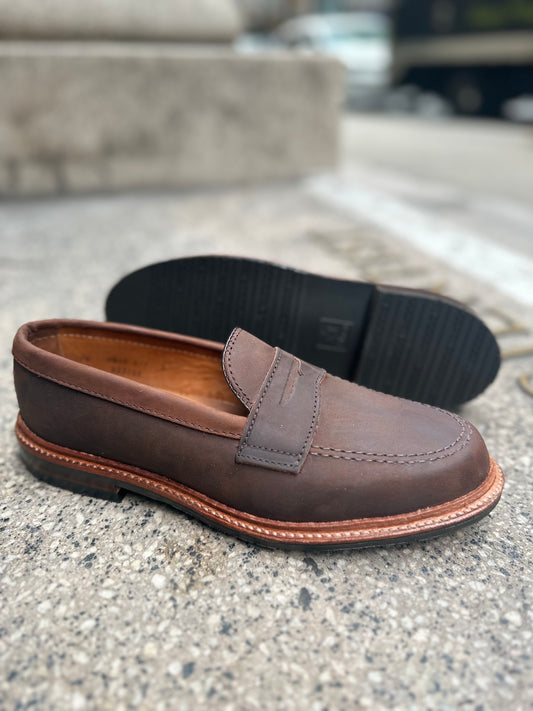 The Alden Madison D2216L - Smooth Tobacco Chamois Penny Loafer, with antique edge and visible stitching on a rubber sole, stands upright on concrete while another lies sideways, revealing its intricate tread pattern.
