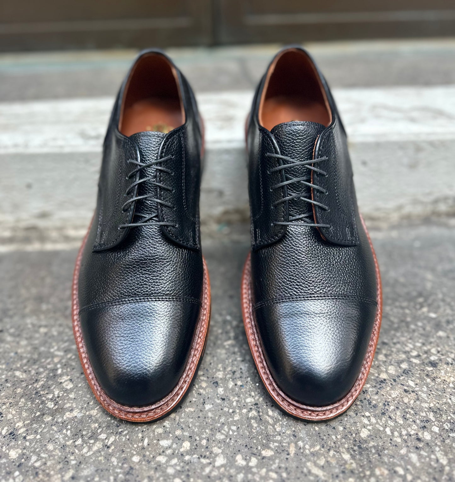 A pair of Alden Madison 22826C Black JS Grain "Derosa" Blucher dress shoes with brown soles and classic laces, shown side by side on rough concrete near a step.