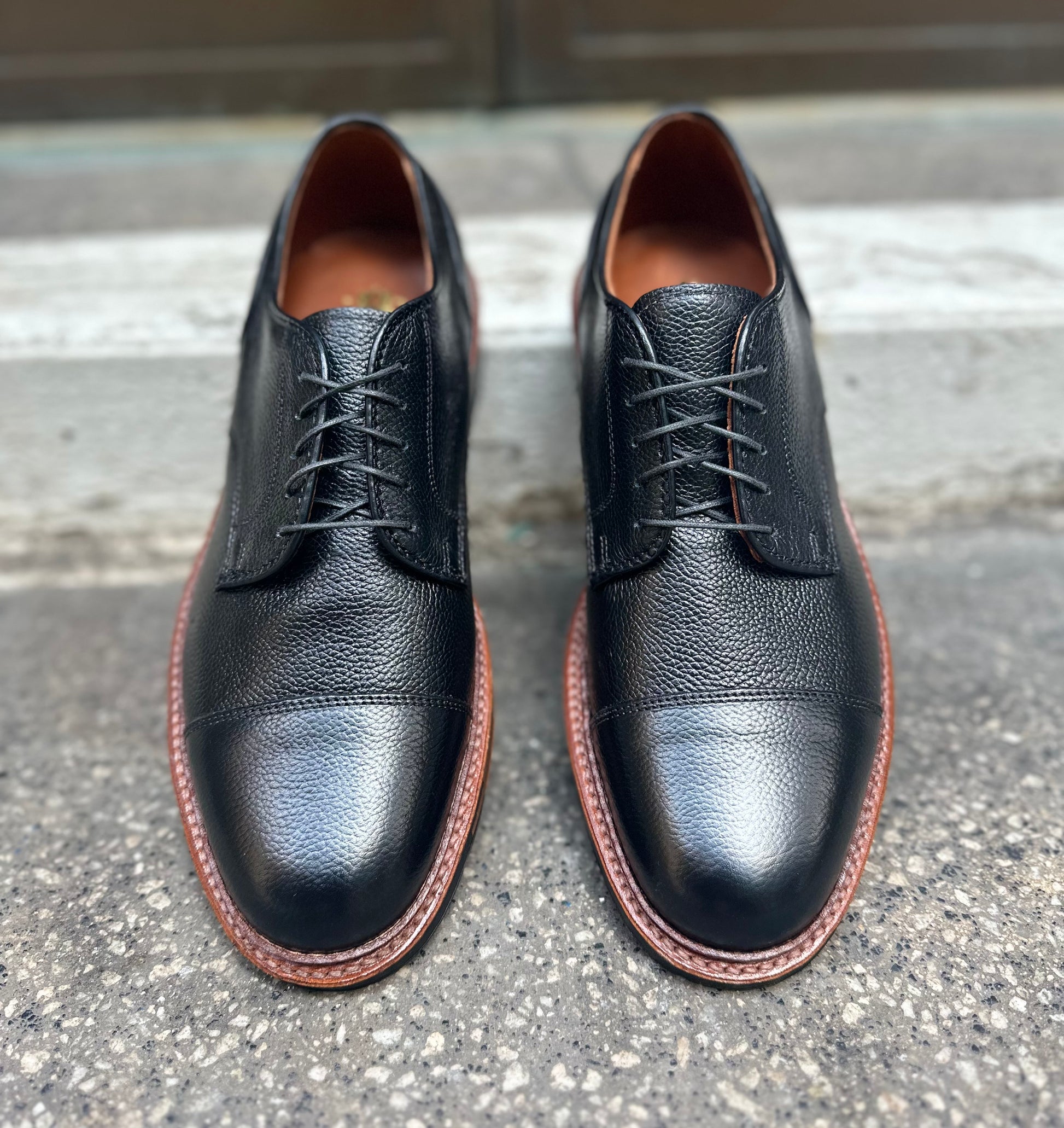 A pair of Alden Madison 22826C Black JS Grain "Derosa" Blucher dress shoes with brown soles and classic laces, shown side by side on rough concrete near a step.
