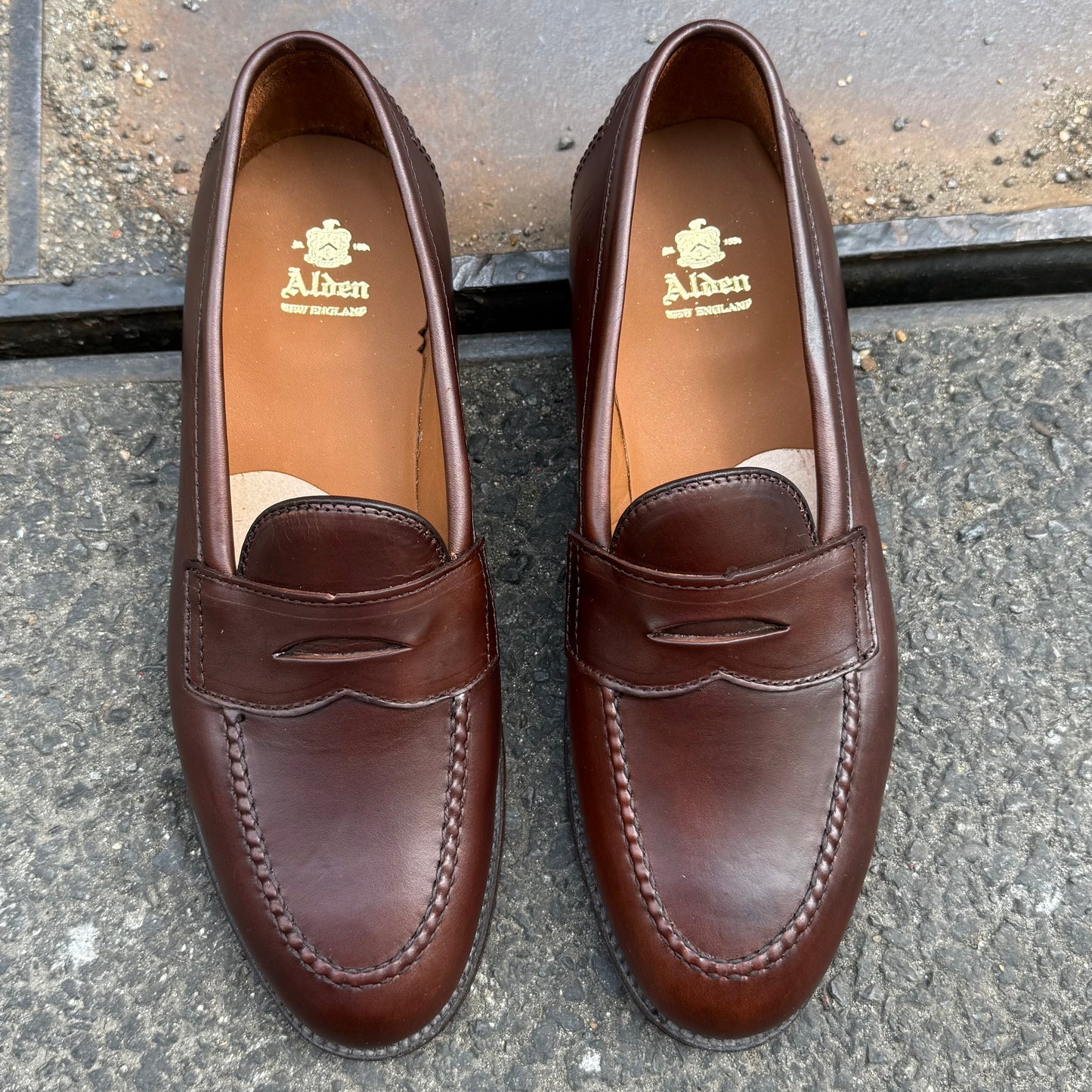 A pair of Alden Madison D5211 Brown CXL Flex Penny Loafers is side by side on rough gray pavement, featuring visible stitching, Brown Chromexcel leather, a leather sole, and the Alden logo in gold on the insoles.