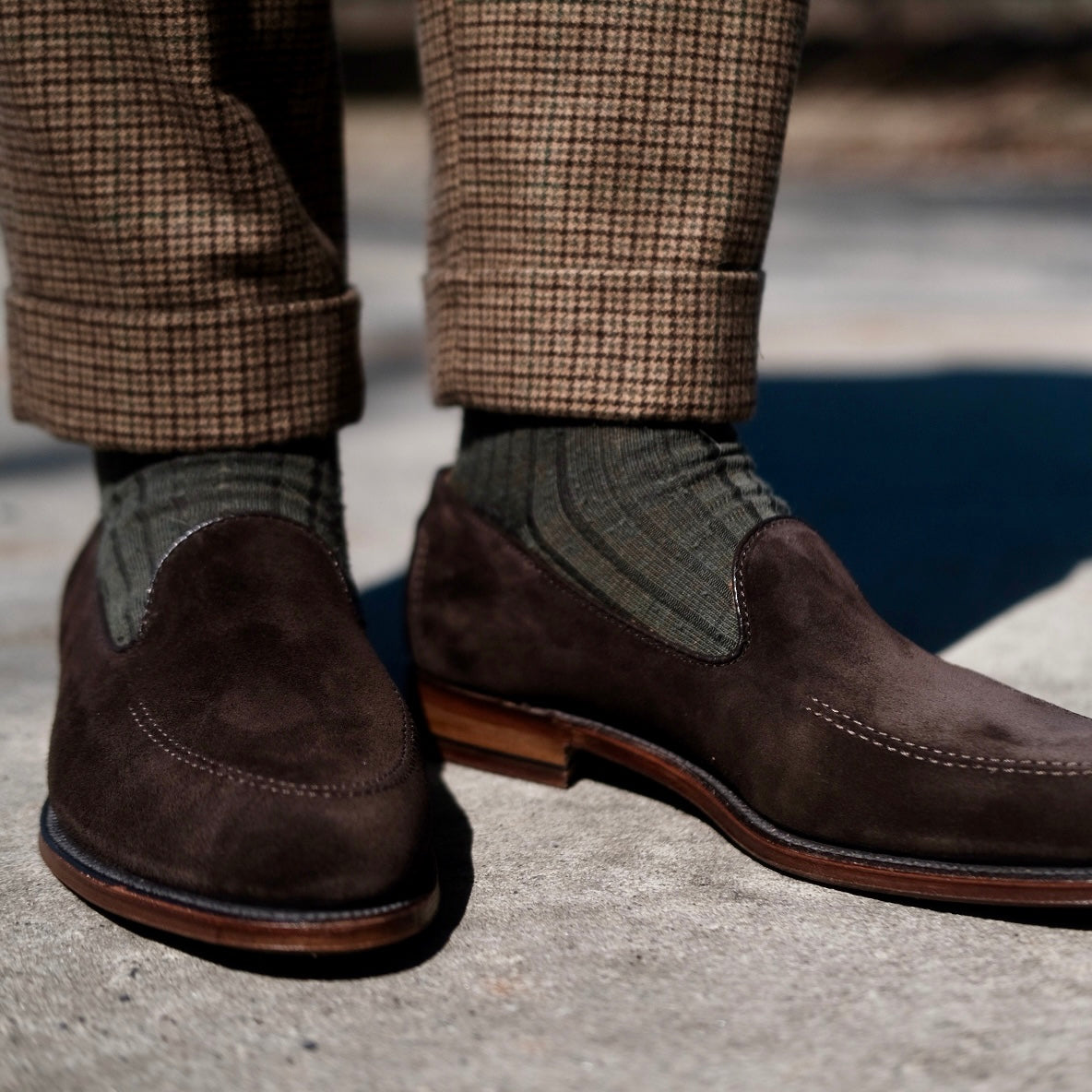 Close-up of a person wearing the ALDEN SHOES D5105 - Vanderbilt in Dark Brown Suede, featuring a Flex Leather Sole, styled with olive green ribbed socks and brown plaid pants, standing on a sunlit concrete surface.