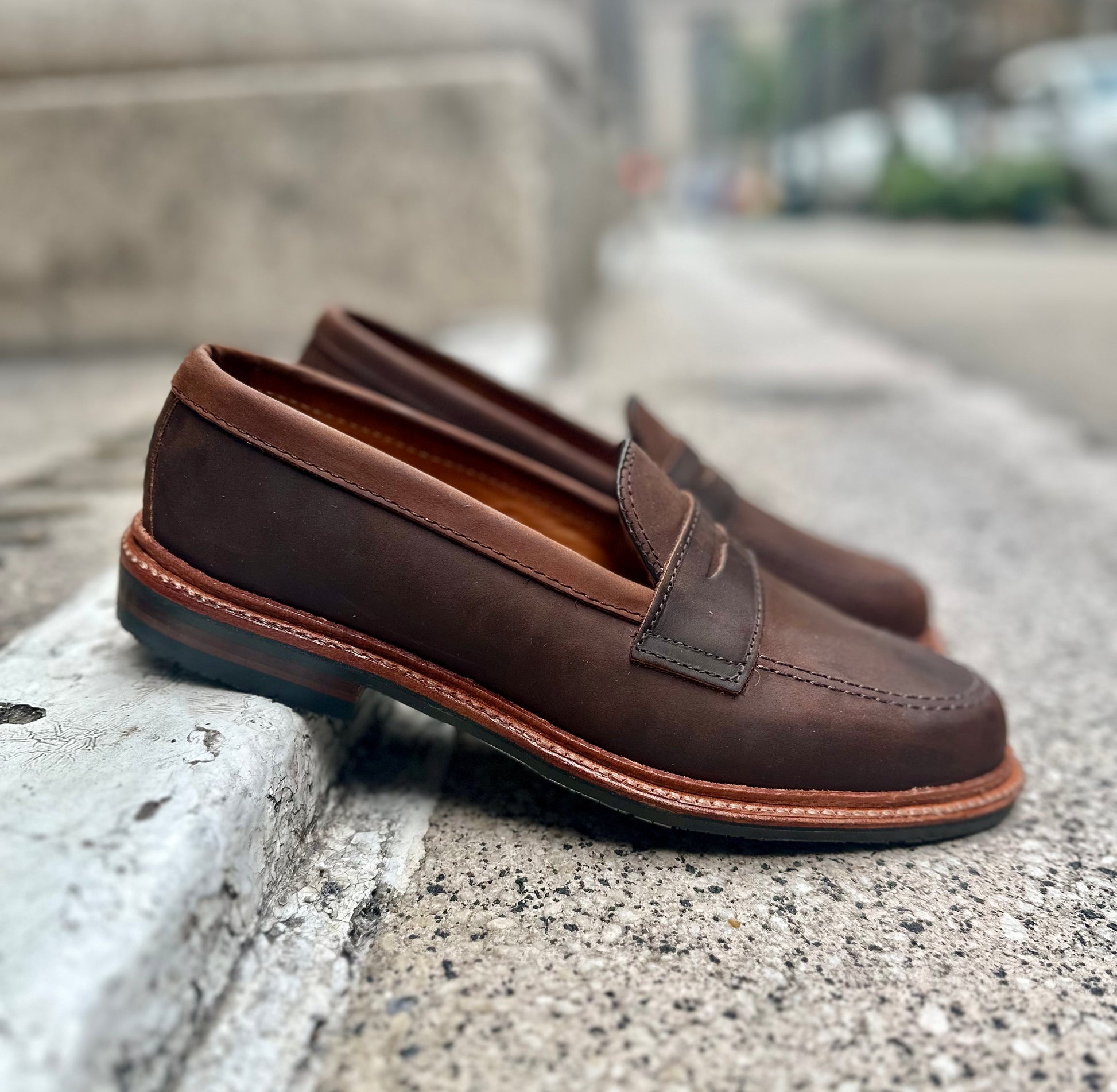 The Alden Madison D2216L - Smooth Tobacco Chamois Penny Loafers, featuring an antique edge and visible sole, sit on a concrete surface with a slightly blurred urban backdrop that highlights their stitch detailing and tobacco smooth finish.