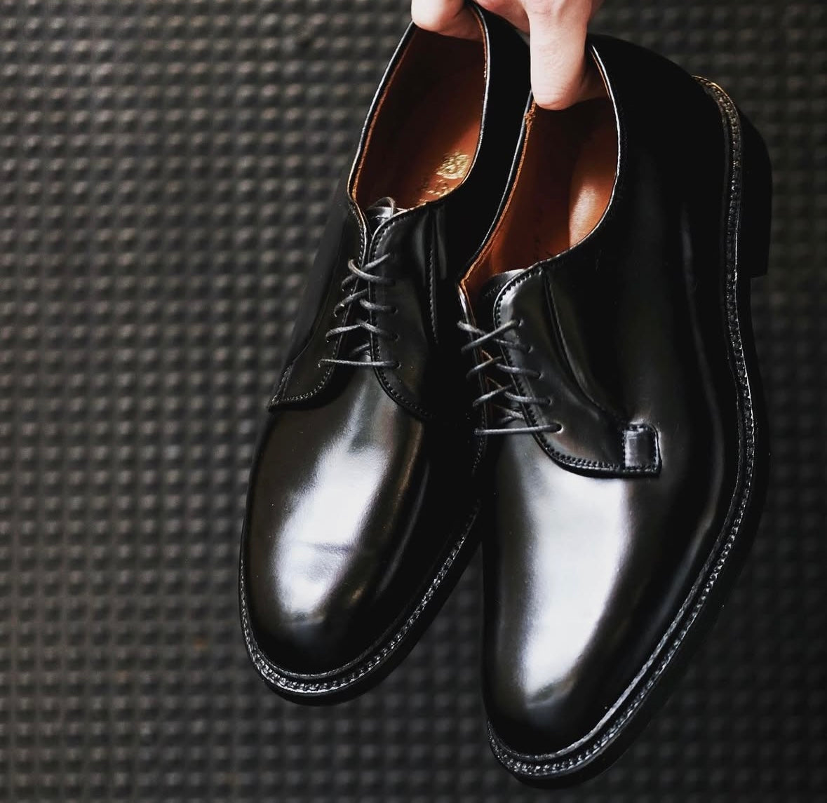 A person holds a pair of Alden Madison 9901 - Plain Toe Blucher Black in Shell Cordovan dress shoes with laces and Double Oak Leather Sole, set against a dark textured background.