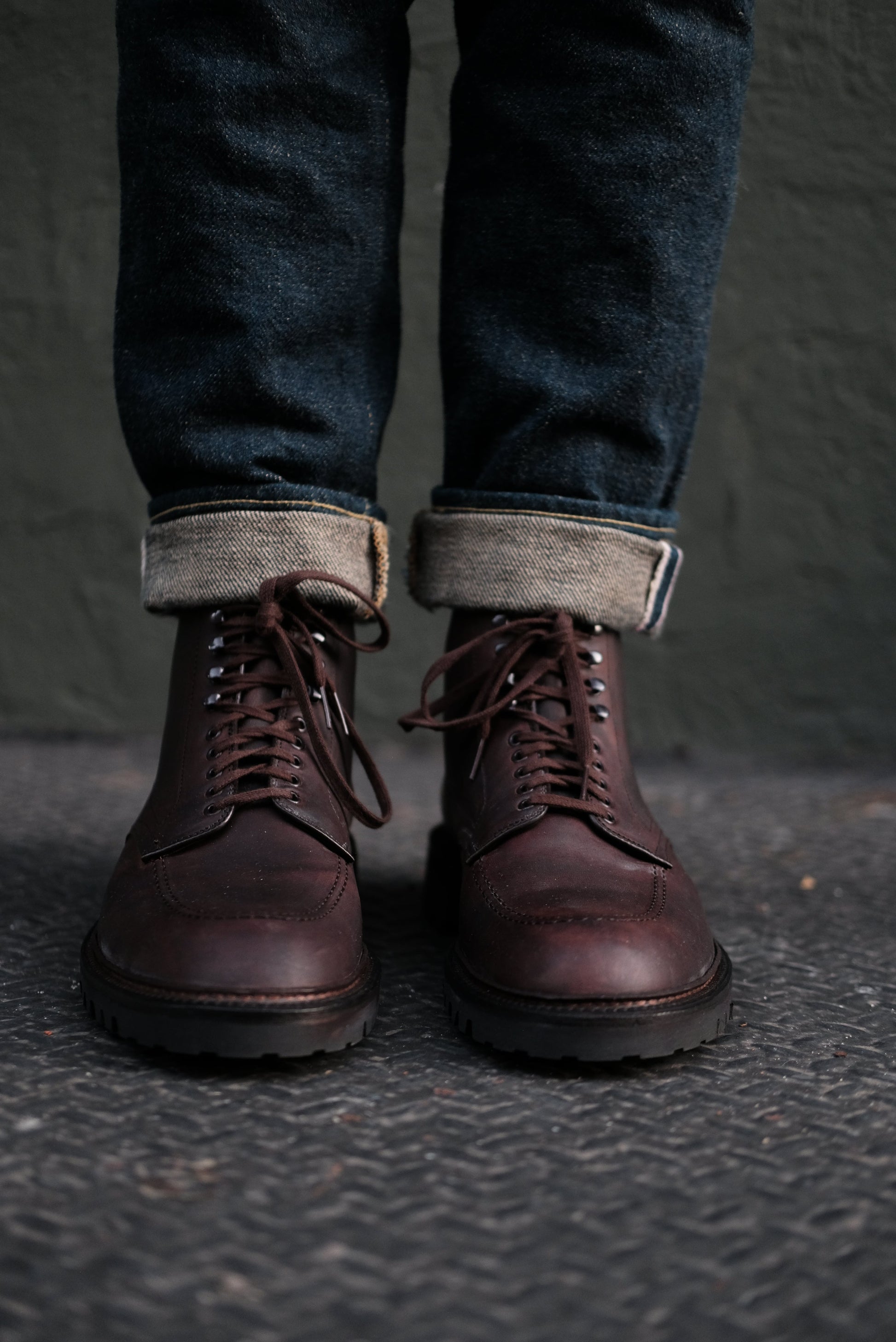 A person in cuffed dark blue jeans and Alden Madisons 404 - Indy Boot in Kudu (Oiled CXL) with a Vibram Lug Outsole stands on a textured surface, set against a plain dark wall.