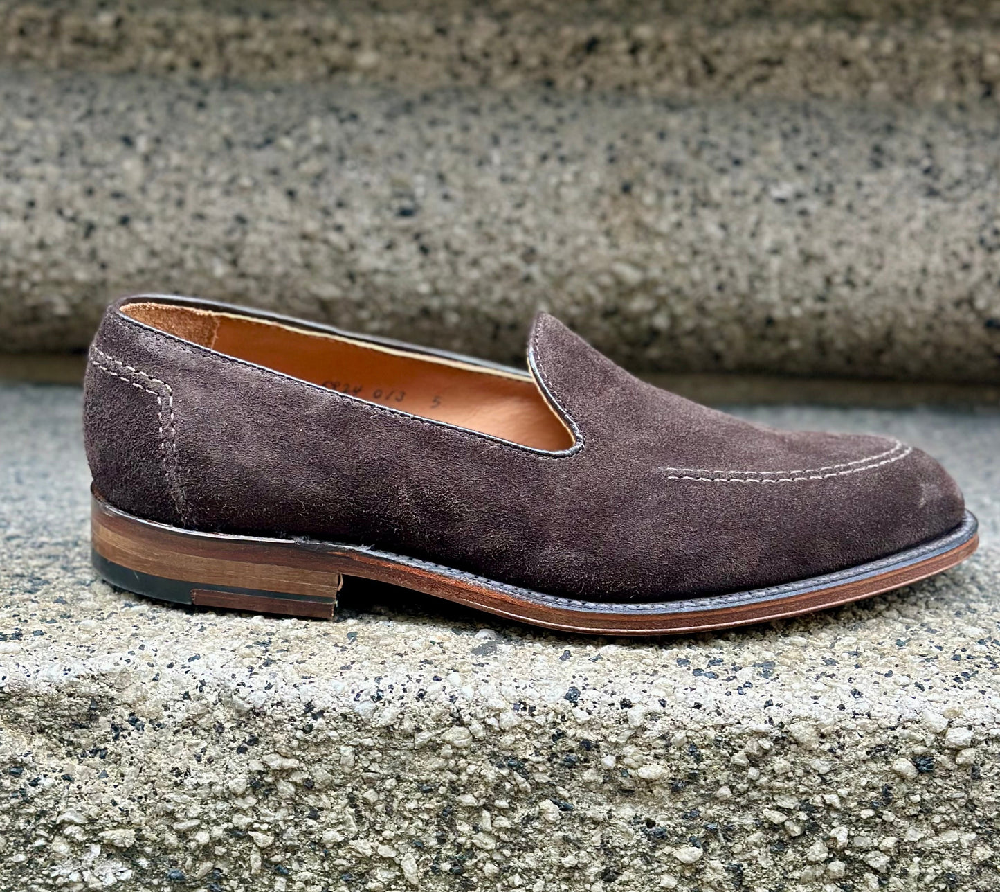 The D5105 - Vanderbilt in Dark Brown Suede by ALDEN SHOES, featuring a Flex Leather Sole and visible stitching, rests elegantly on a rough concrete step.