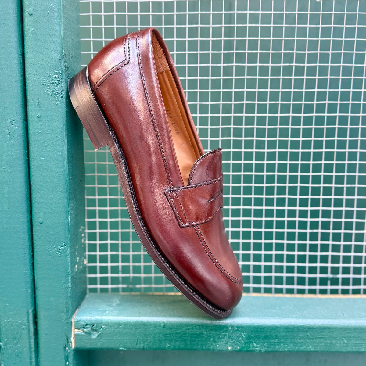 A single Alden Madison D5214 - Brown Calf Flex Penny loafer, expertly crafted from luxurious Brown Vegano Calf on the Copley Last, rests on its side atop a teal-framed wire mesh background, showcasing its visible stitching and sophisticated Flex Leather Sole.