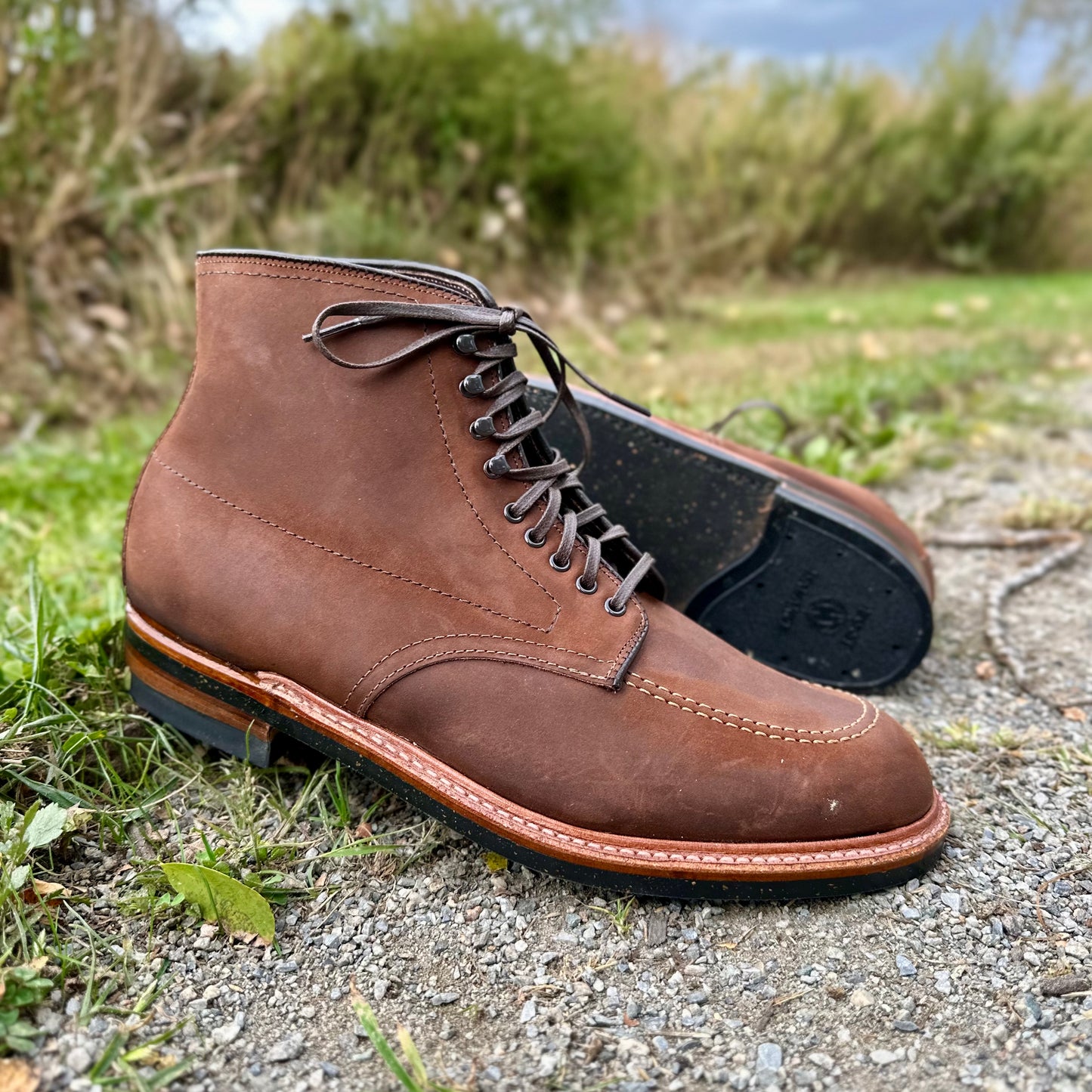 An Alden Madison D2909H Indy Boot in Tobacco Smooth Chamois sits upright on a gravel path, encircled by vibrant greenery. The boots, with dark soles and brown laces, include one standing while the other lies elegantly on its side.