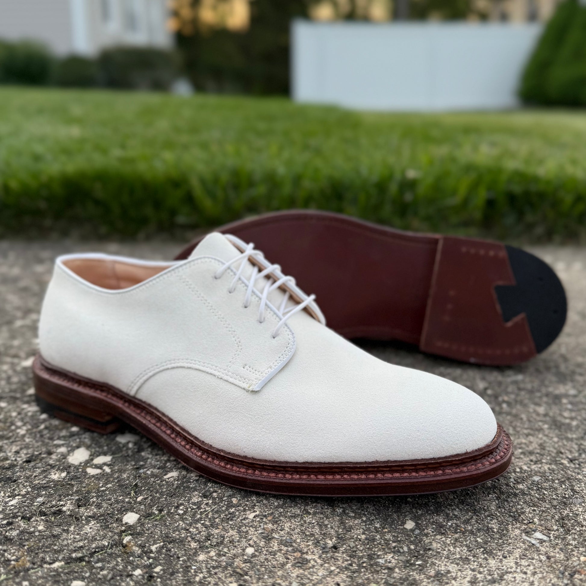 The Alden Madison 29330F - Unlined Dover in Ivory Suede with brown leather soles, crafted on the Barrie last, is displayed outdoors—one upright, one on its side—on textured concrete with grass in the background.