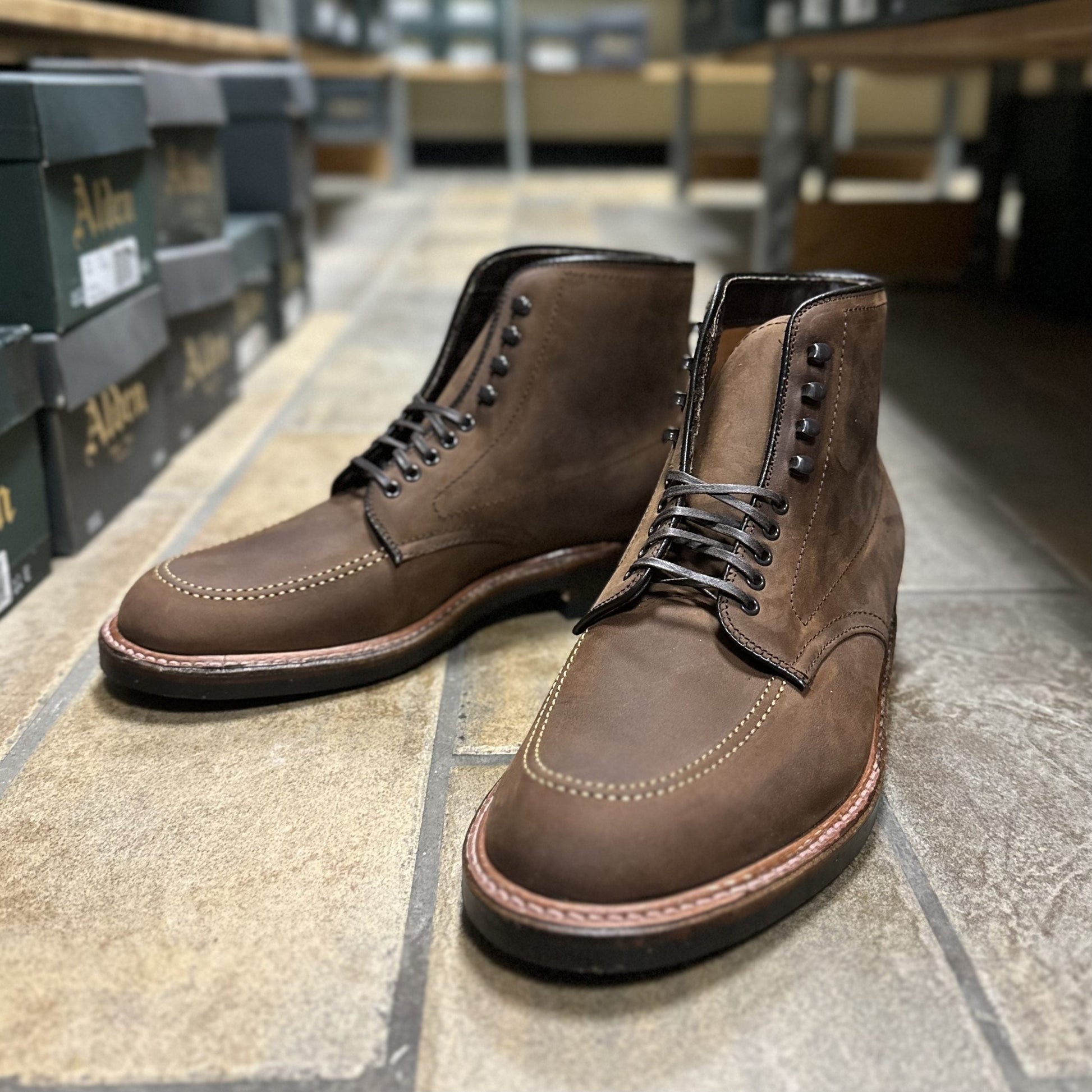 A pair of Alden Madisons D2909H Indy Boots in Tobacco Smooth Chamois with dark eyelets and stitching detail, crafted on a Trubalance Last, rests elegantly on a tiled floor, while shoe boxes sit neatly on shelves in the background.